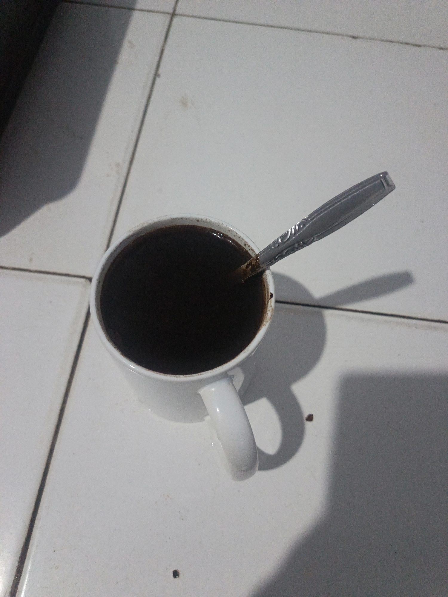 A white ceramic mug filled with dark liquid, presumably coffee, sits on a tiled floor. A silver spoon is partially submerged in the liquid, angled slightly upwards. The mug has a visible handle. The floor tiles are white and appear to be laid in a grid pattern with visible grout lines. Shadows are cast on the floor, suggesting overhead lighting. The scene is viewed from a top-down perspective. The image does not contain any identifiable landmarks or contextual elements that would place it specifically within the city of Gorontalo, Indonesia.