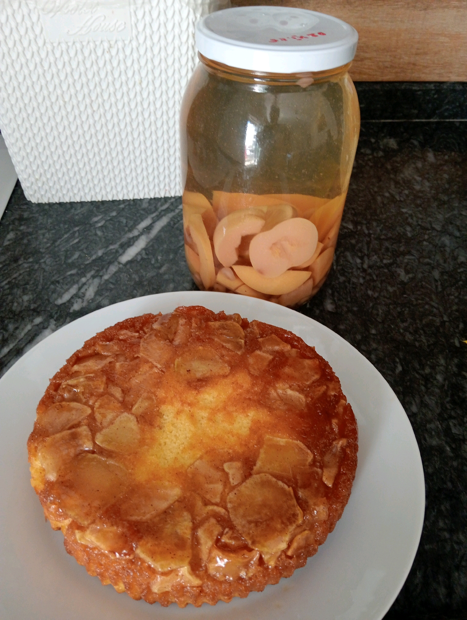 A close-up, high-angle shot displays a round, golden-brown cake with visible pieces of fruit on a white plate. To the upper right of the cake is a clear glass jar filled with sliced, light-colored fruit in a clear liquid, with a white lid. In the background, to the left, is a white woven basket with raised lettering. The scene is set on a dark, speckled countertop. The location is an indoor kitchen in Silifke, Turkey.