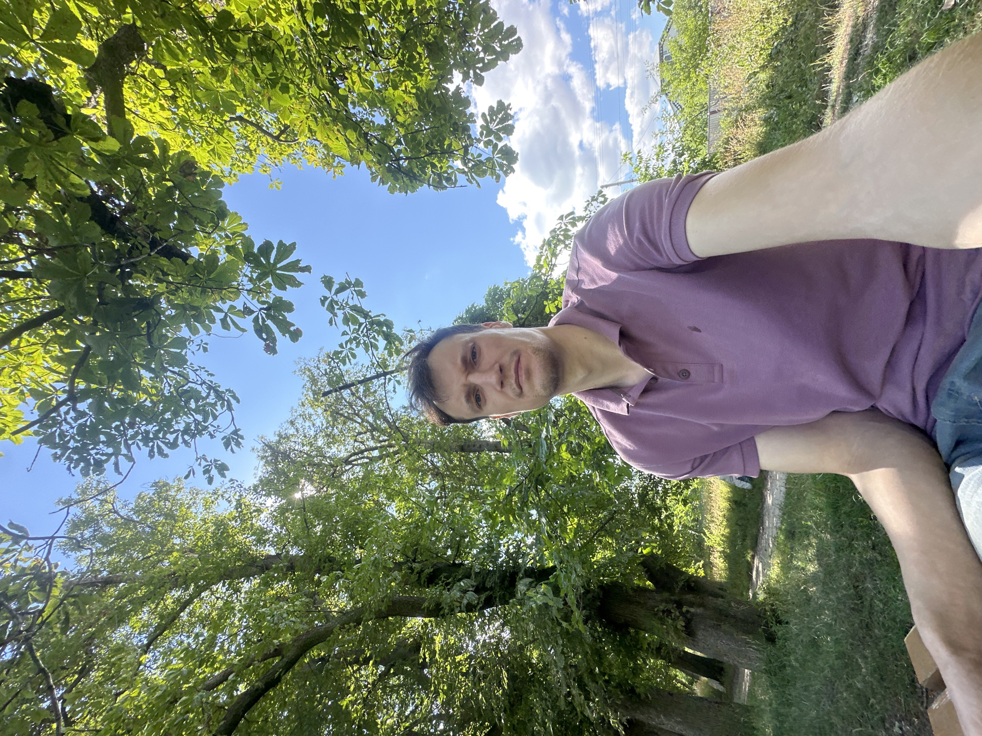 The original image shows a young adult male, appearing to be in his late 20s or early 30s, taking a selfie outdoors. He is wearing a purple polo shirt and blue jeans. The man has short brown hair and is looking directly at the camera with a neutral expression. He is seated on what appears to be a park bench, with his right arm extended as if holding the camera.
The background is a lush green park or garden with large trees. The leaves of the trees are vibrant green, suggesting it is a warm season. Sunlight is filtering through the branches, creating dappled light and shadows on the ground. The sky is a clear blue with a few fluffy white clouds. In the distance, there are hints of residential buildings and a fence, suggesting the park may be near a populated area.
The overall impression is a casual, outdoor portrait taken on a sunny day. The presence of trees and foliage suggests a natural setting within a city or town. Without specific landmarks, pinpointing the exact location within Makiv, Ukraine is not possible from the visual information alone.