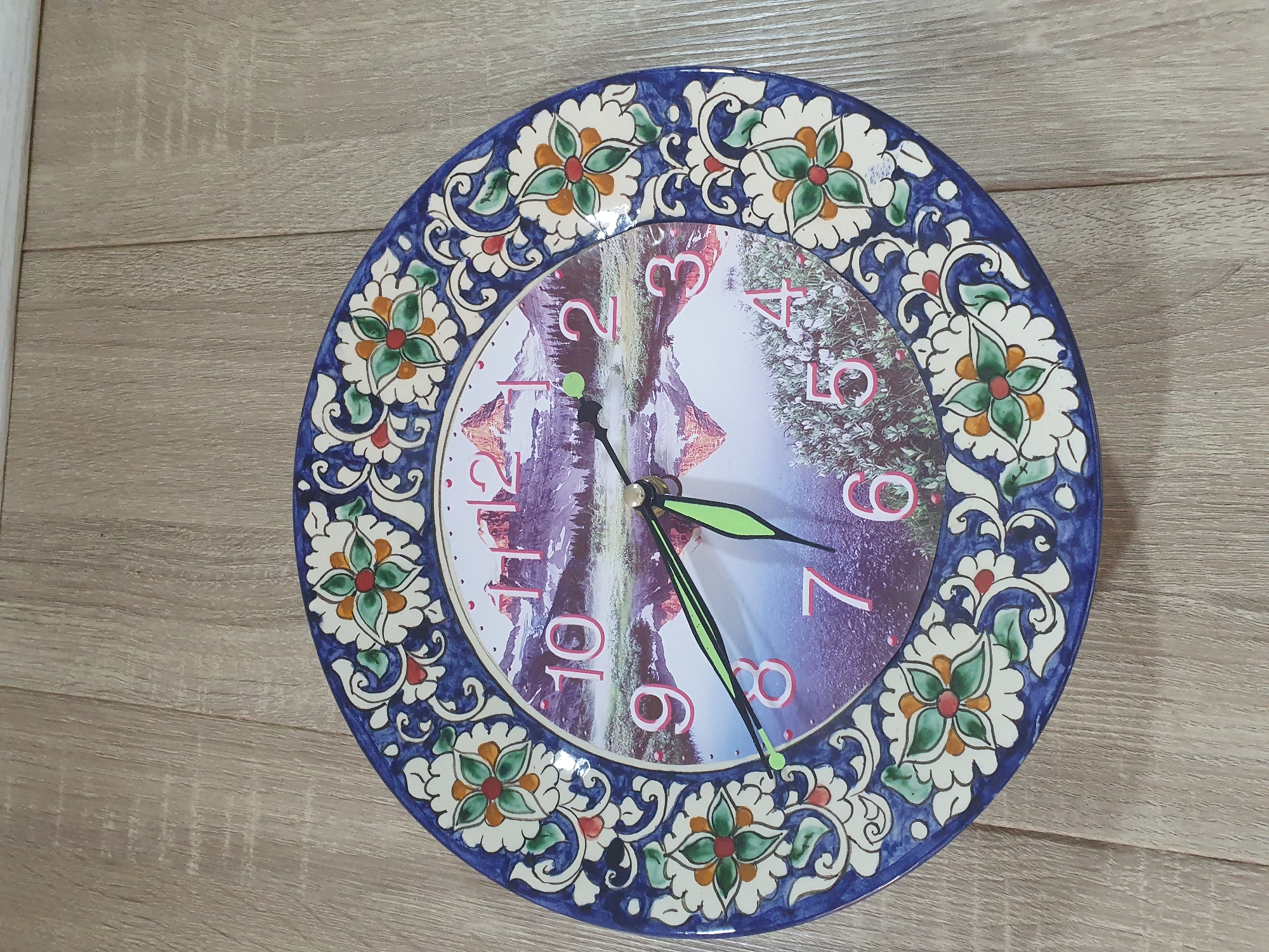 A decorative wall clock with a scenic landscape image is displayed on a wooden surface. The clock face features a picture of a waterfall cascading down rocky cliffs with a hint of trees on the right side. The numbers on the clock are in a vibrant red, with the numbers 2, 3, 4, 5, 6, 7, 8, 9, 10, and 11 clearly visible. The clock hands are a bright green, with the hour hand pointing towards the 2 and the minute hand pointing towards the 5. The clock has a decorative blue border with floral motifs in white, green, and orange. The background is a light-colored wooden planked surface, suggesting an indoor setting. The image does not contain any people, and no specific time of day, weather conditions, or emotions can be determined. The location context is Tashkent, Uzbekistan.