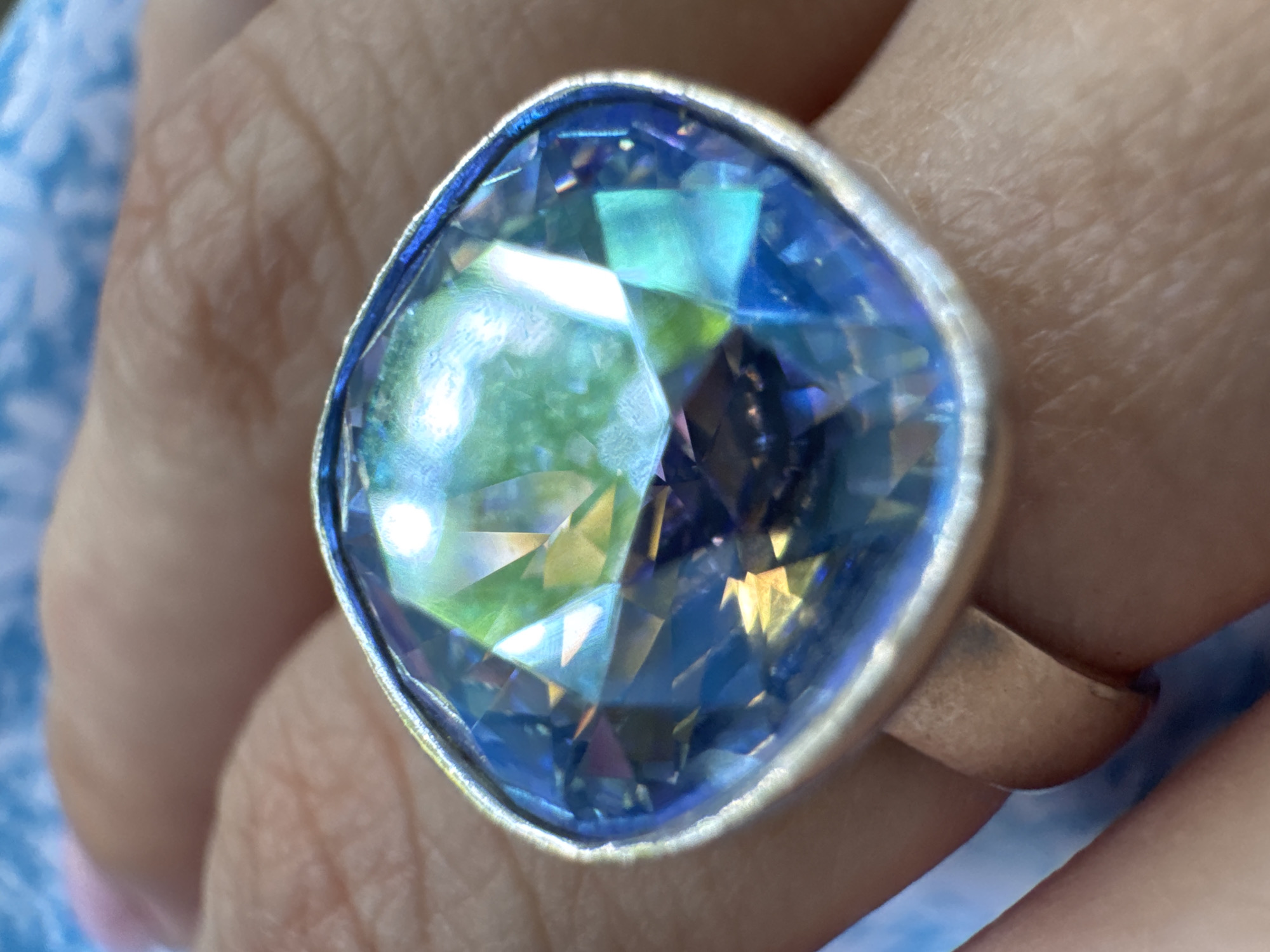 A close-up shot focuses on a person's hand wearing a ring with a large, multifaceted blue gemstone. The gemstone catches the light, displaying various shades of blue, green, and hints of yellow. The ring itself appears to be made of silver or a similar metal. The hand is resting on a surface with a light blue, possibly fabric, pattern, and the background is softly blurred, suggesting an indoor setting or an outdoor scene with shallow depth of field. The lighting is bright, creating reflections on the gemstone, and the time of day is likely daytime. There is no visible text in the image.