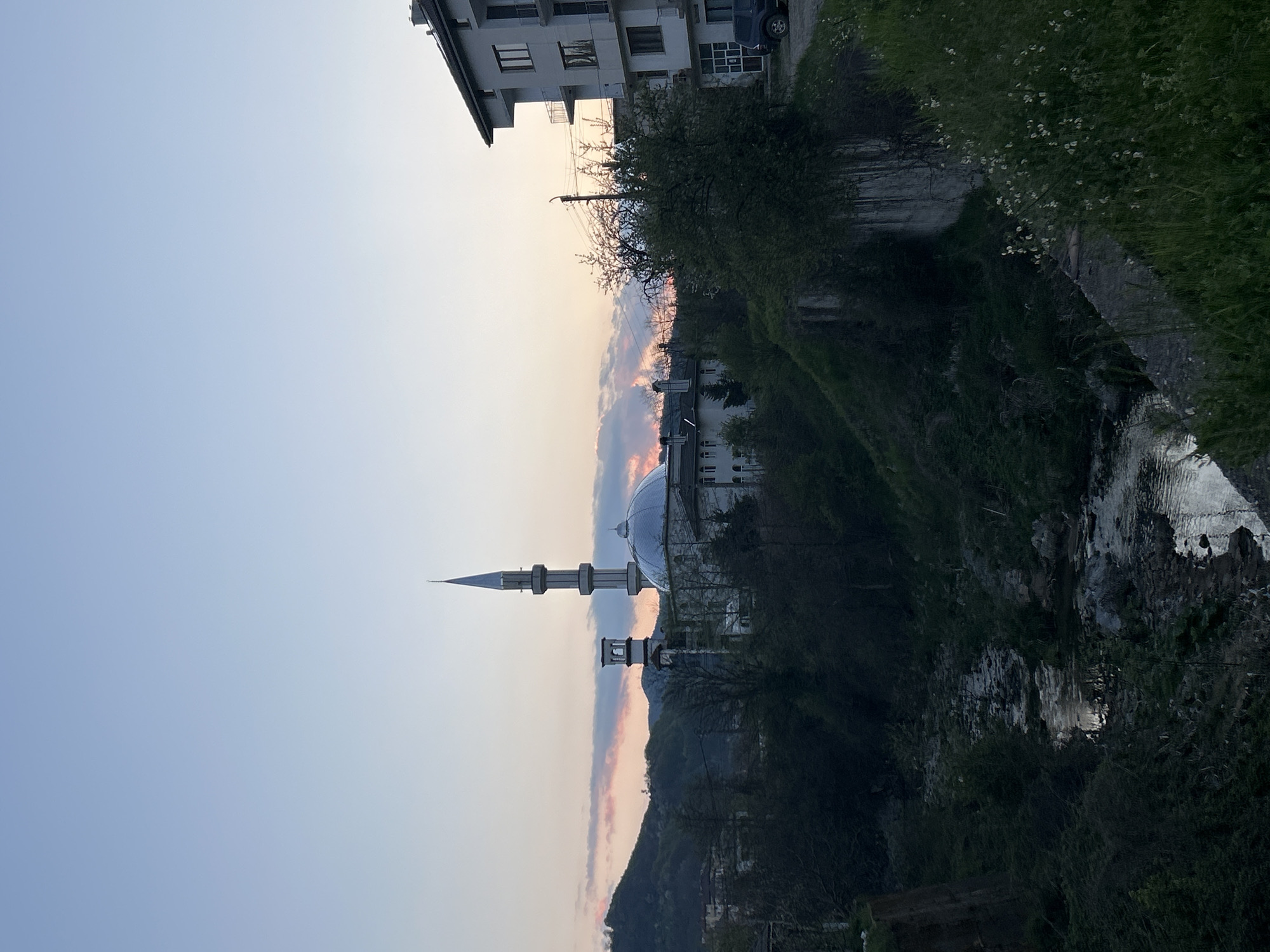 A landscape-oriented shot captures a mosque in Zlatograd, Bulgaria. The mosque is characterized by a large, metallic dome and a slender minaret. Adjacent to the mosque, on the right, flows a narrow riverbank with dense vegetation. A residential building with multiple floors stands at the top left corner of the frame. The backdrop features a twilight sky with hues of pink and blue.