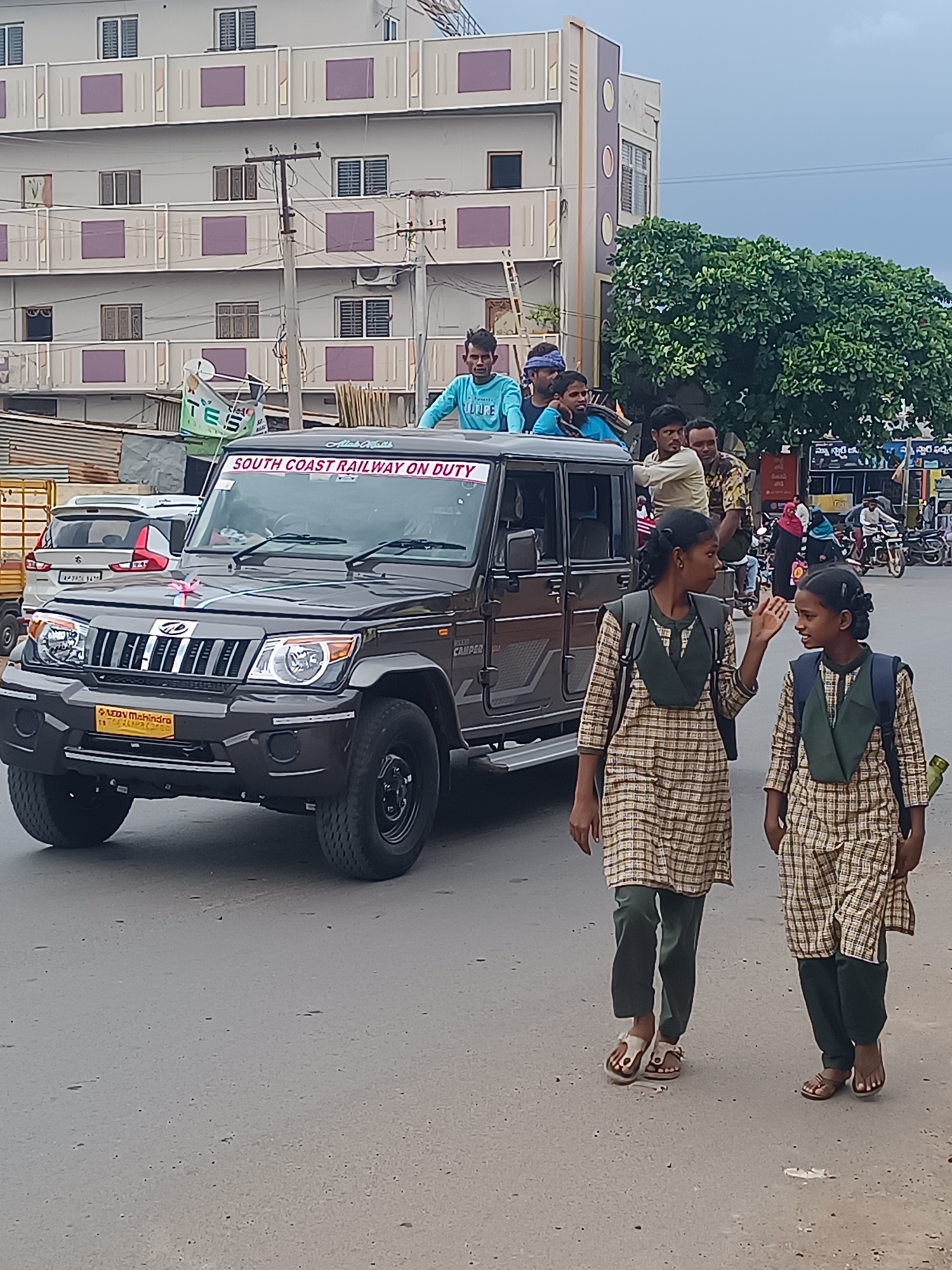 In Bethamcherla, India, a dark-colored Mahindra Bolero Camper is parked on a paved road, adorned with a pink ribbon on its hood, possibly for a ceremony or event. The vehicle's windshield displays a prominent red banner that reads "SOUTH COAST RAILWAY ON DUTY". Several men are positioned inside and on top of the vehicle. Two young girls, dressed in plaid school uniforms and carrying backpacks, walk on the side of the road. One girl gestures with her right hand while walking. In the background, a multi-story building with purple and white accents stands tall, alongside a large green tree. The sky is overcast. Other vehicles, including a white SUV, are visible on the road, along with motorcycles and a few pedestrians further down the street.