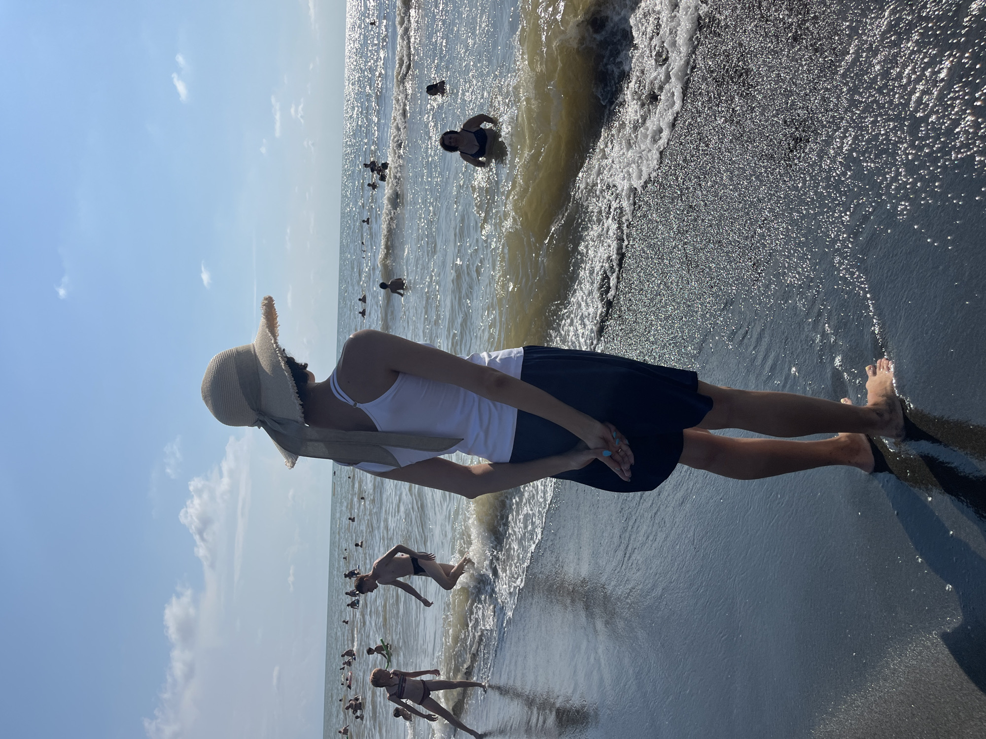 The image depicts a sunny day at the Ureki beach in Georgia, characterized by its black magnetic sand and the Black Sea. In the foreground, a person wearing a straw hat, white tank top, and dark skirt stands facing away from the camera, their hands clasped behind their back. The person is standing at the edge of the water, with their feet partially submerged in the shallow waves.
Further into the sea, numerous individuals are visible engaging in various water activities. Some are swimming, while others are standing or wading in the water. The sea is moderately wavy, with sunlight glinting off the surface. The background shows a clear blue sky with scattered white clouds. The overall scene conveys a sense of a popular summer destination with people enjoying the beach and the sea.