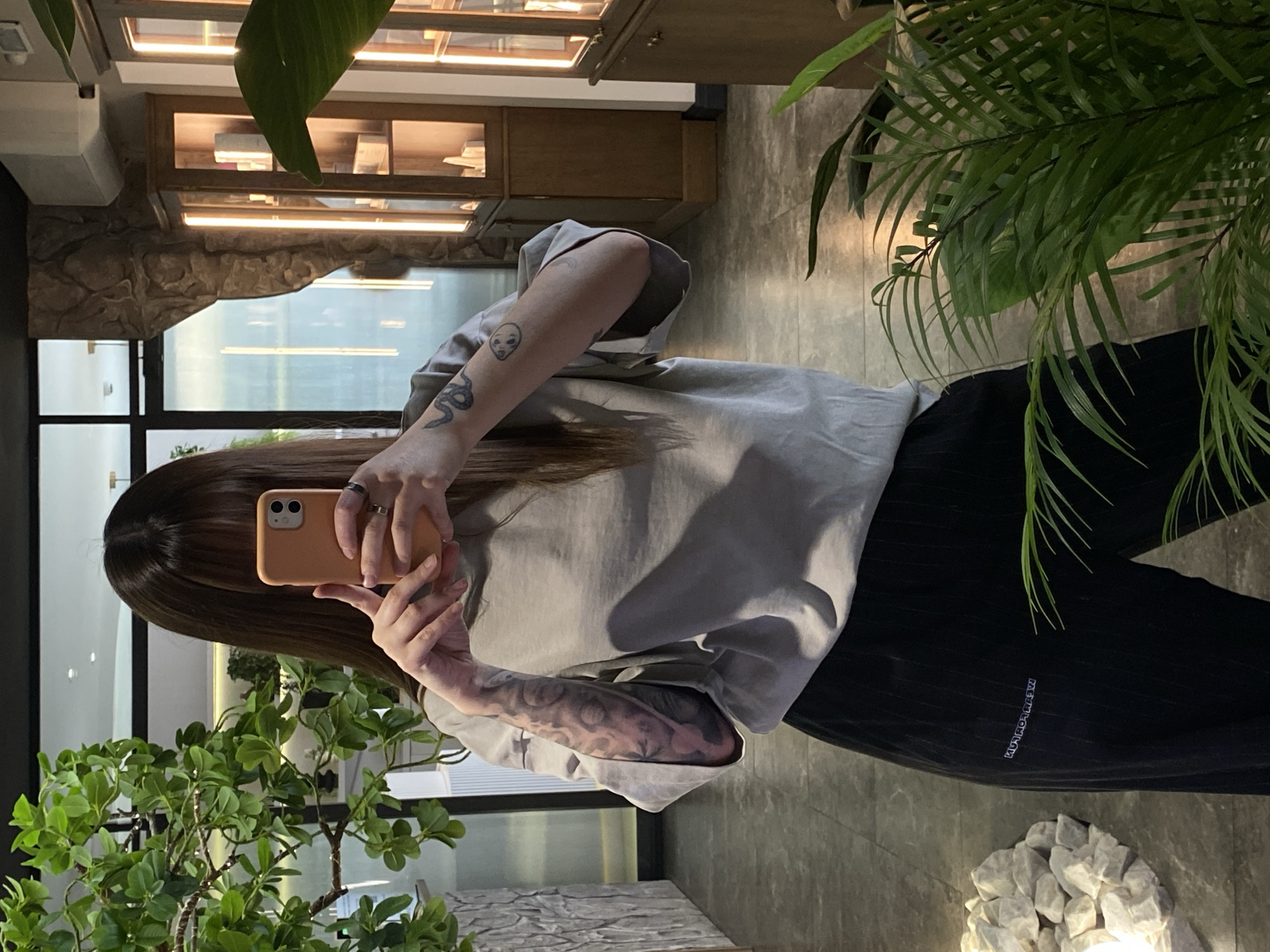 A person is holding an orange smartphone and taking a selfie in an interior space. The person has long brown hair and is wearing a light grey oversized t-shirt and black pants. The person has tattoos on their arms. There are plants on either side of the person, and a display case with items inside in the background. The floor appears to be made of grey tile. The person appears to be indoors in Votkinsk, Russia.