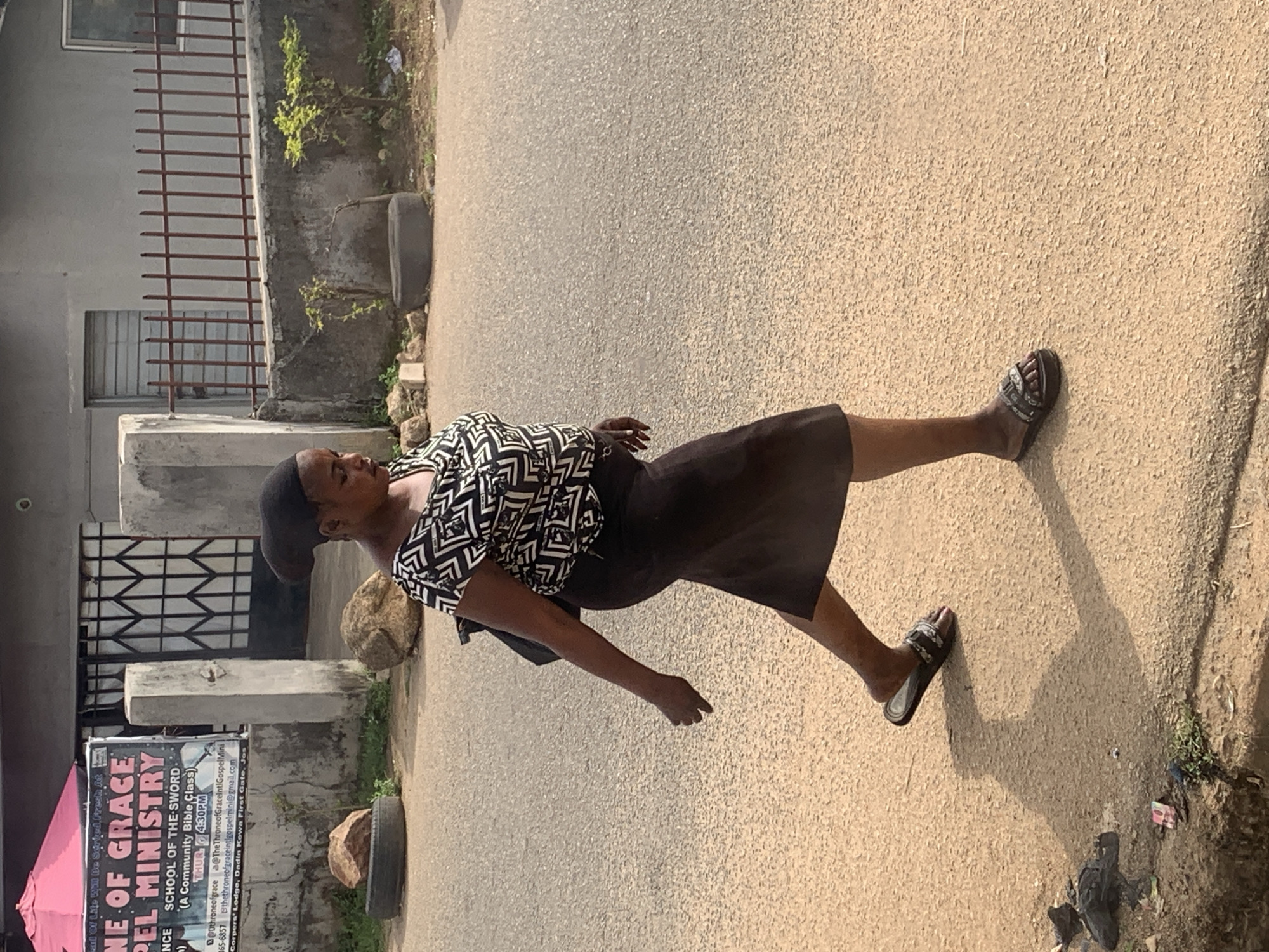 An outdoor, sunlit street scene shows a woman walking on a paved road. The woman is of dark complexion, wearing a black head covering, a black and white patterned short-sleeved blouse, a black knee-length skirt, and dark-colored sandals. She appears to be looking towards her left.
The ground is a light-colored, pebbled asphalt road or pathway. Strong shadows are cast on the ground, indicating bright daylight, likely during the mid-morning or early afternoon.
To the woman's left (viewer's right, if upright) is a building with a simple architectural style, featuring a metal grille or fence and a window. Concrete structures and some greenery are also visible near the building. To her right (viewer's left), there is a large banner or sign that reads "THRONE OF GRACE GOSPEL MINISTRY" and advertises "SCHOOL OF THE SWORD. (A Community Bible Class)" held on "THUR 4:30PM." Contact details such as "@Dthroneofgrace," "@TheThroneofGraceintGospelMini," a phone number "07066165-6857," and an email "thethroneofgraceintgospelmini@gmail.com" are present. The sign also indicates the location as "Corpers' Lodge, Dadin Kowa First Gate, Jos." A pink umbrella or canopy is partially visible near the bottom of the sign. Several rocks and a discarded tire are visible on the side of the road.