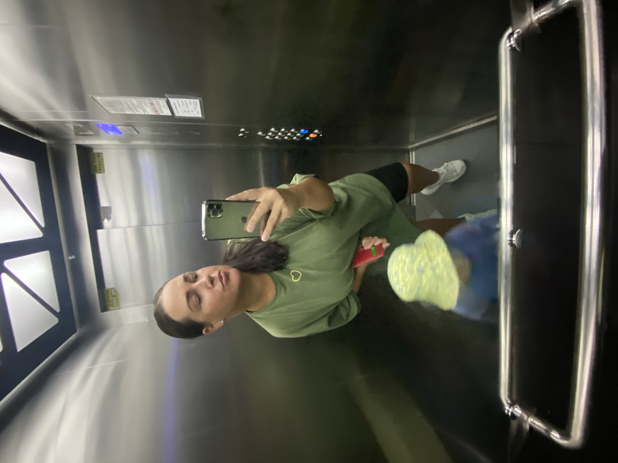 A woman, wearing an olive green t-shirt and black shorts, takes a selfie in an elevator. She is holding a smartphone in her right hand. A child, wearing a blue shirt and a yellow and blue patterned sun hat, is visible in the reflection of the elevator doors. The elevator has stainless steel walls and a control panel with buttons. The scene is set in an elevator, likely in Sochi, Russia.