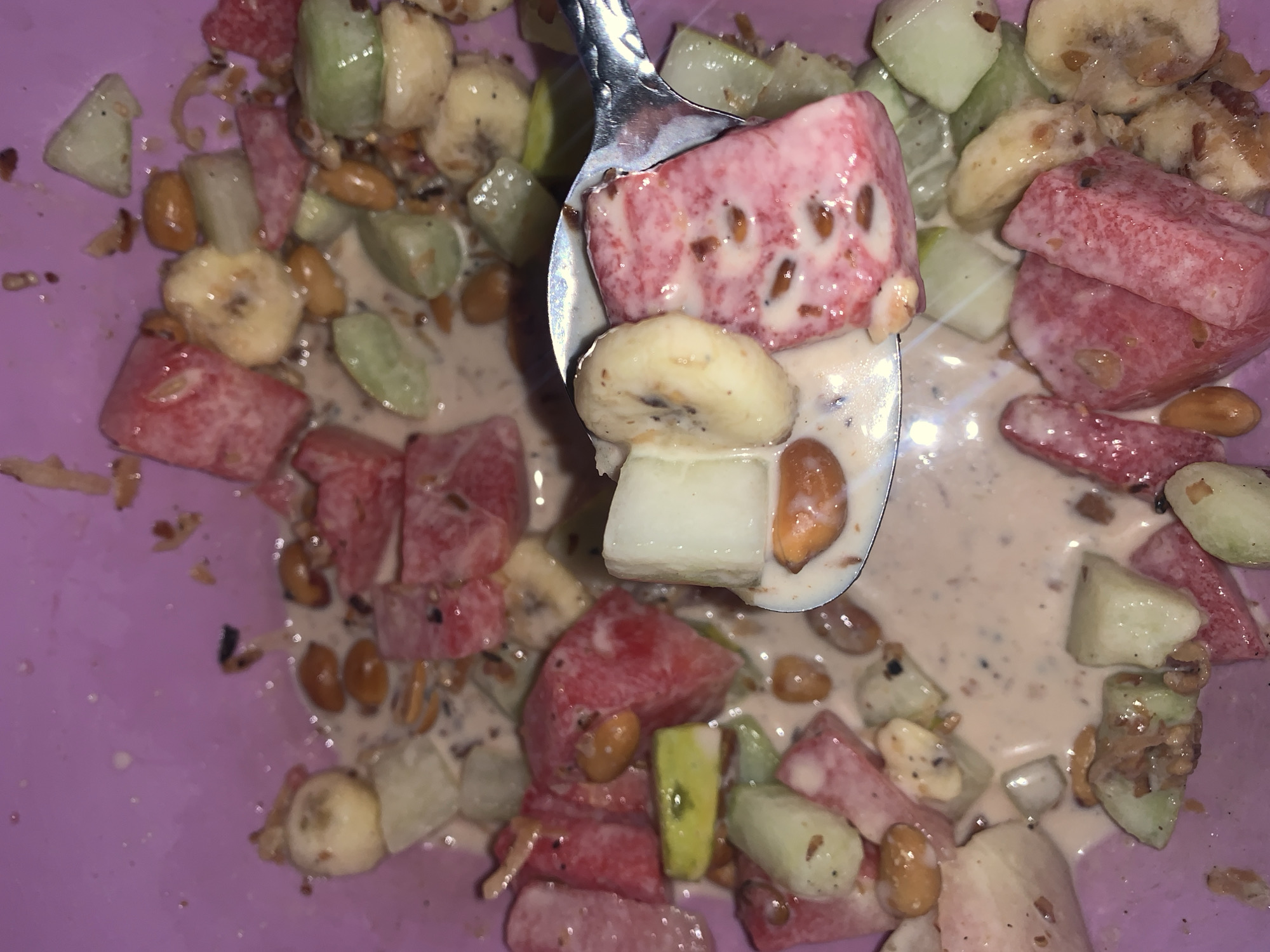 An overhead shot captures a vibrant fruit salad served in a lavender-colored bowl. The bowl is filled with a mix of chopped fruits and other ingredients, coated in a light beige, creamy dressing.
A silver spoon is prominent in the center, scooping up a portion of the salad. On the spoon, a large piece of red watermelon with visible black seeds, a slice of banana, and a piece of a light green fruit or vegetable (possibly cucumber or unripe apple) are visible, all coated in the same creamy sauce.
Within the bowl, the fruit salad consists primarily of cuboid chunks of bright red watermelon, some showing black seeds. Sliced bananas, identifiable by their characteristic shape and occasional small brown spots, are also interspersed throughout. Small, light green pieces, similar to those on the spoon, add another color and texture. Numerous light brown peanuts are mixed in, adding a nutty element. Dark specks, possibly ground spices like pepper or cinnamon, are visible on many of the fruit pieces and in the creamy dressing.
The overall impression is a fresh, colorful, and refreshing dish, likely a snack or dessert. The creamy coating suggests a yogurt-based or sweetened dressing. There is no visible text in the image. The lighting appears to be indoor artificial light, casting some reflections on the spoon and wet surfaces of the fruit.