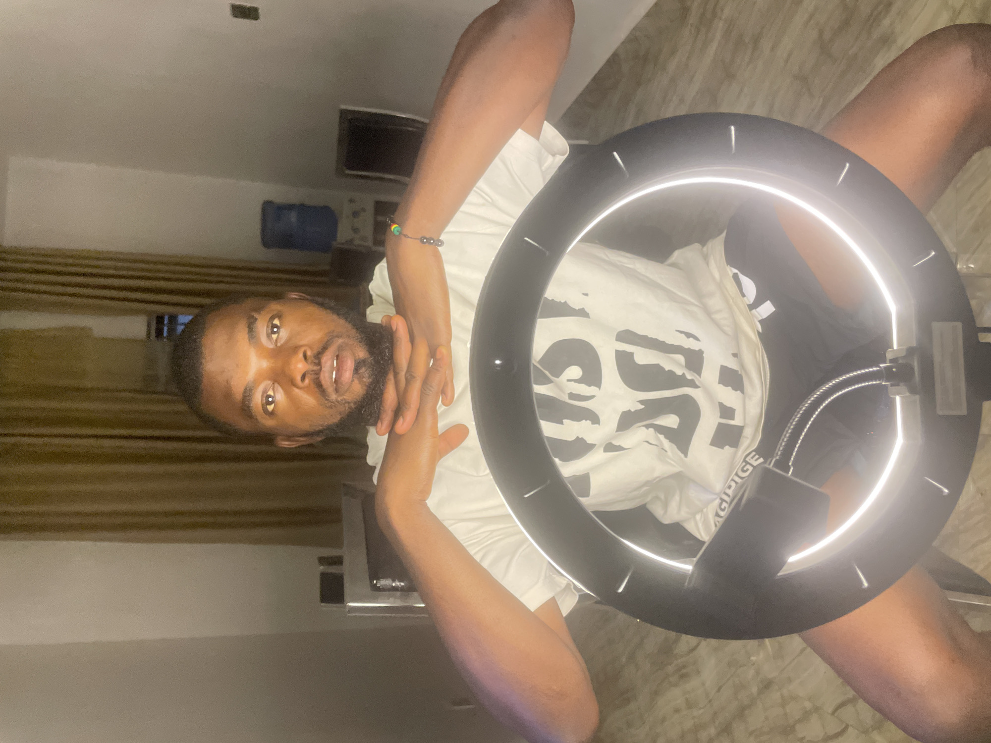 A man is posing in front of a ring light. He has dark skin, a beard, and is wearing a white t-shirt. His hands are raised near his face. He appears to be indoors with curtains and furniture visible in the background. The location in the city of Jos, Nigeria is not discernible from the image.