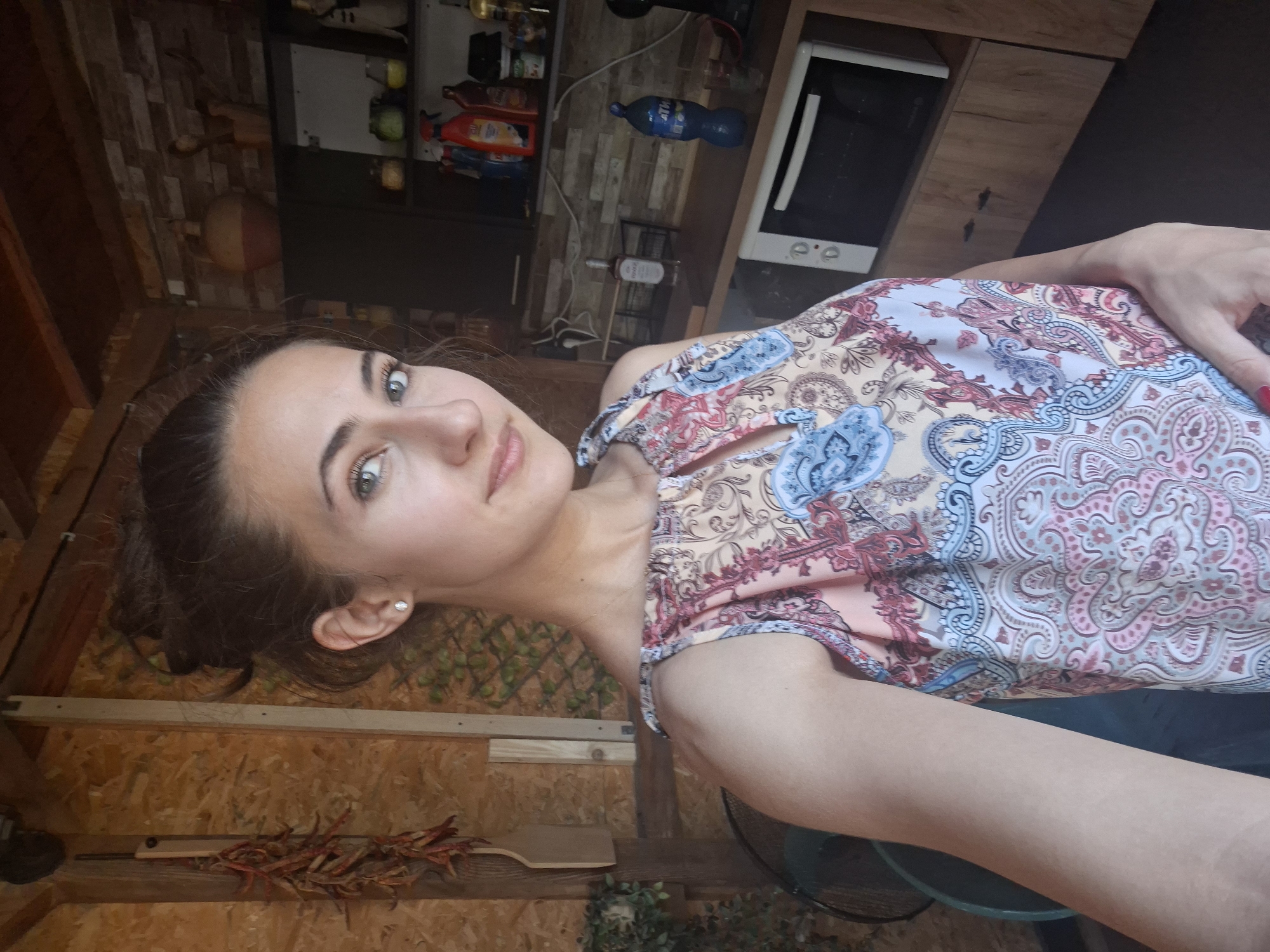 A young woman with brown hair pulled up in a bun and green eyes smiles into the camera, her head slightly tilted. She is wearing a sleeveless top with a colorful paisley print in shades of pink, blue, and beige. A small diamond stud earring is visible in her left ear. Her right arm is extended towards the camera. The background appears to be an outdoor kitchen area. To her left, a string of dried red peppers hangs on a wooden wall. To her right, shelves are stocked with various bottles and jars, and a small microwave oven is visible. The overall setting suggests a casual, domestic environment.