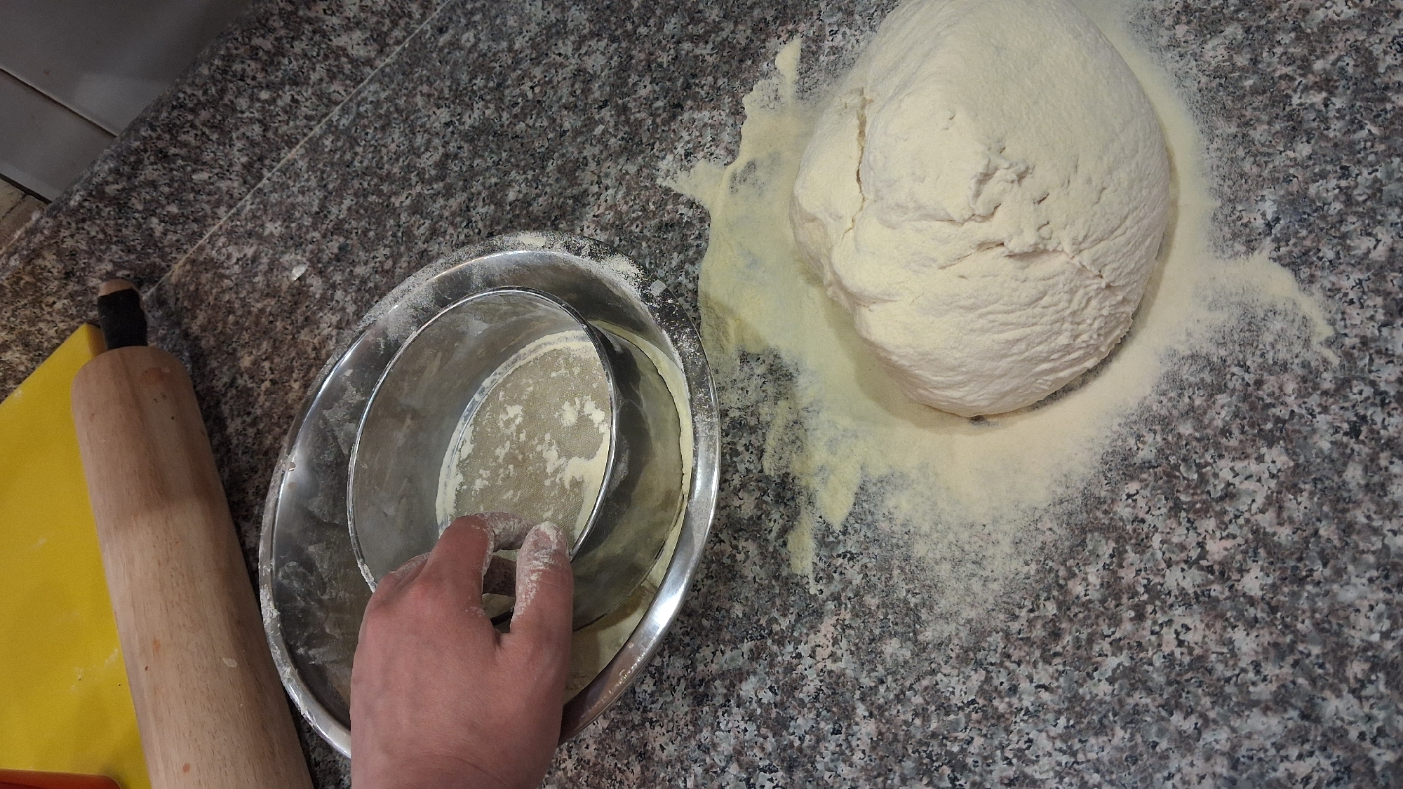 A high-angle shot shows a section of a speckled gray countertop, likely in a kitchen. To the right, a ball of dough is sitting amidst a scattering of flour. To the left, a silver bowl contains a metal sifter dusted with flour, with a hand visible inside the bowl. A wooden rolling pin rests on a yellow cutting board next to the bowl. The city and country (Goce Delcev, Bulgaria) are not evident in the image, so that part of the prompt cannot be fulfilled.