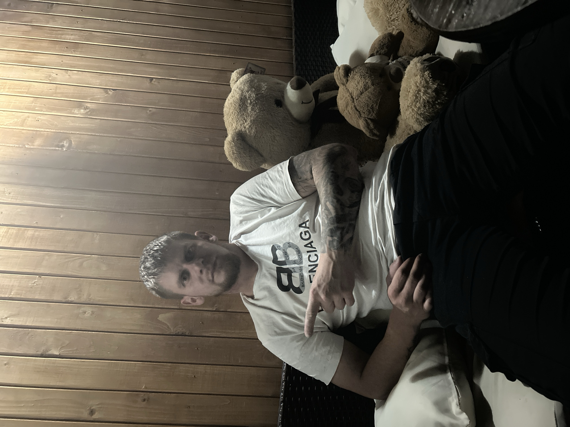 An adult male is seated, facing forward with a slight turn to the right, in a dimly lit, casual setting. He has short, light-colored hair and a short beard. He wears a white short-sleeved t-shirt with a "BALENCIAGA" logo on the chest and dark-colored trousers. Tattoos are visible on his left forearm. His right arm is bent, and his index finger points directly to his left. To his right, on the seating surface, are multiple brown teddy bears, including one large bear prominently positioned next to him and at least two smaller bears below it. The man is seated on a dark-colored piece of furniture, possibly a sofa or lounge chair, with white pillows. The background consists of a vertical wooden plank wall. The location is the city of Unknown, Unknown.