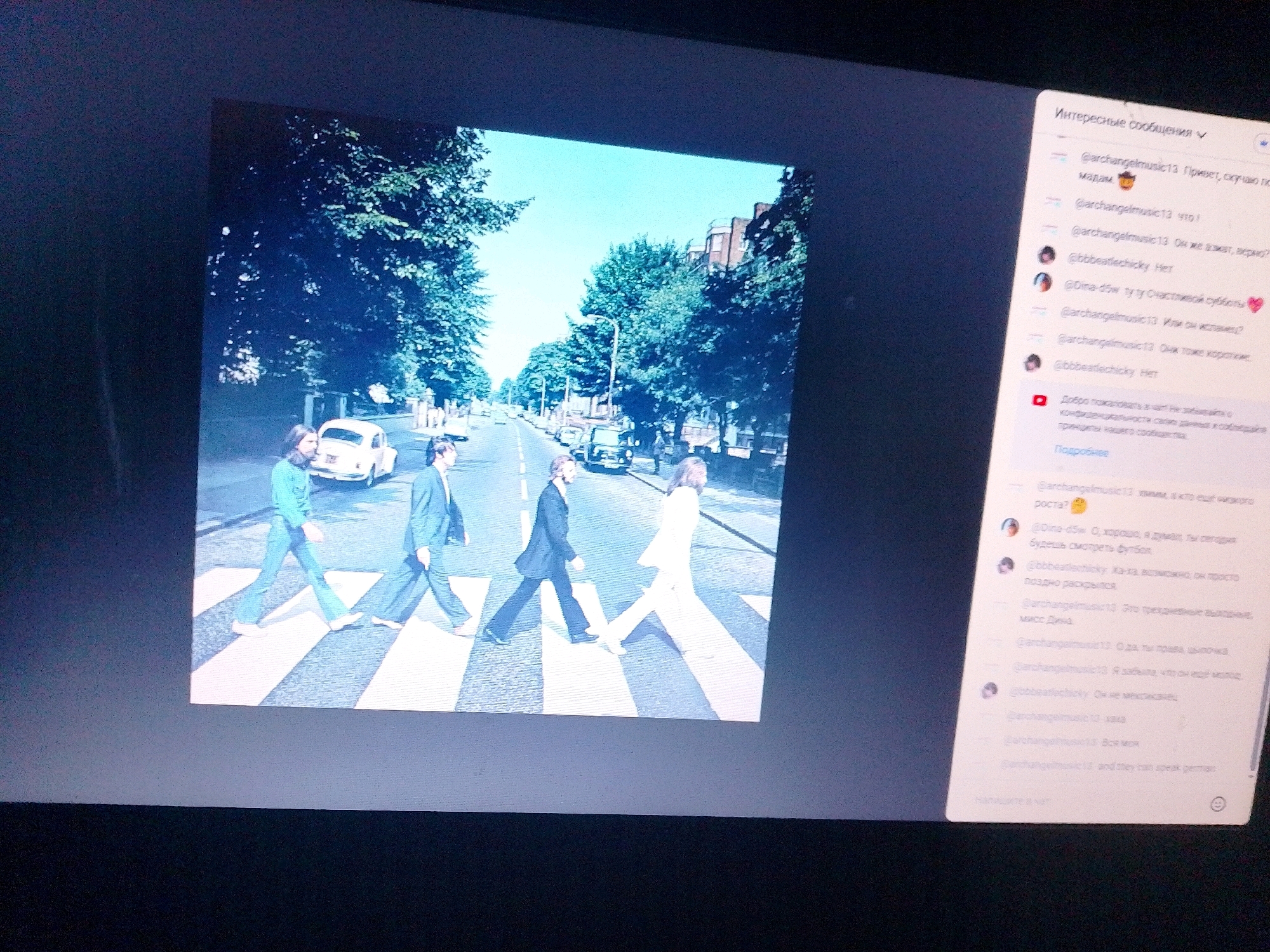The image shows a computer screen displaying a photograph of The Beatles crossing Abbey Road, a famous album cover. To the right of the photograph, a social media comment section is visible. The comments are written in Russian and discuss various topics, including music and personal matters. The photograph depicts four men walking across a pedestrian crossing on a street lined with trees. A white Volkswagen Beetle is parked in the background. The scene is set in a city environment with buildings visible in the distance. There is no information within the image to suggest the location is Myrhorod, Ukraine.