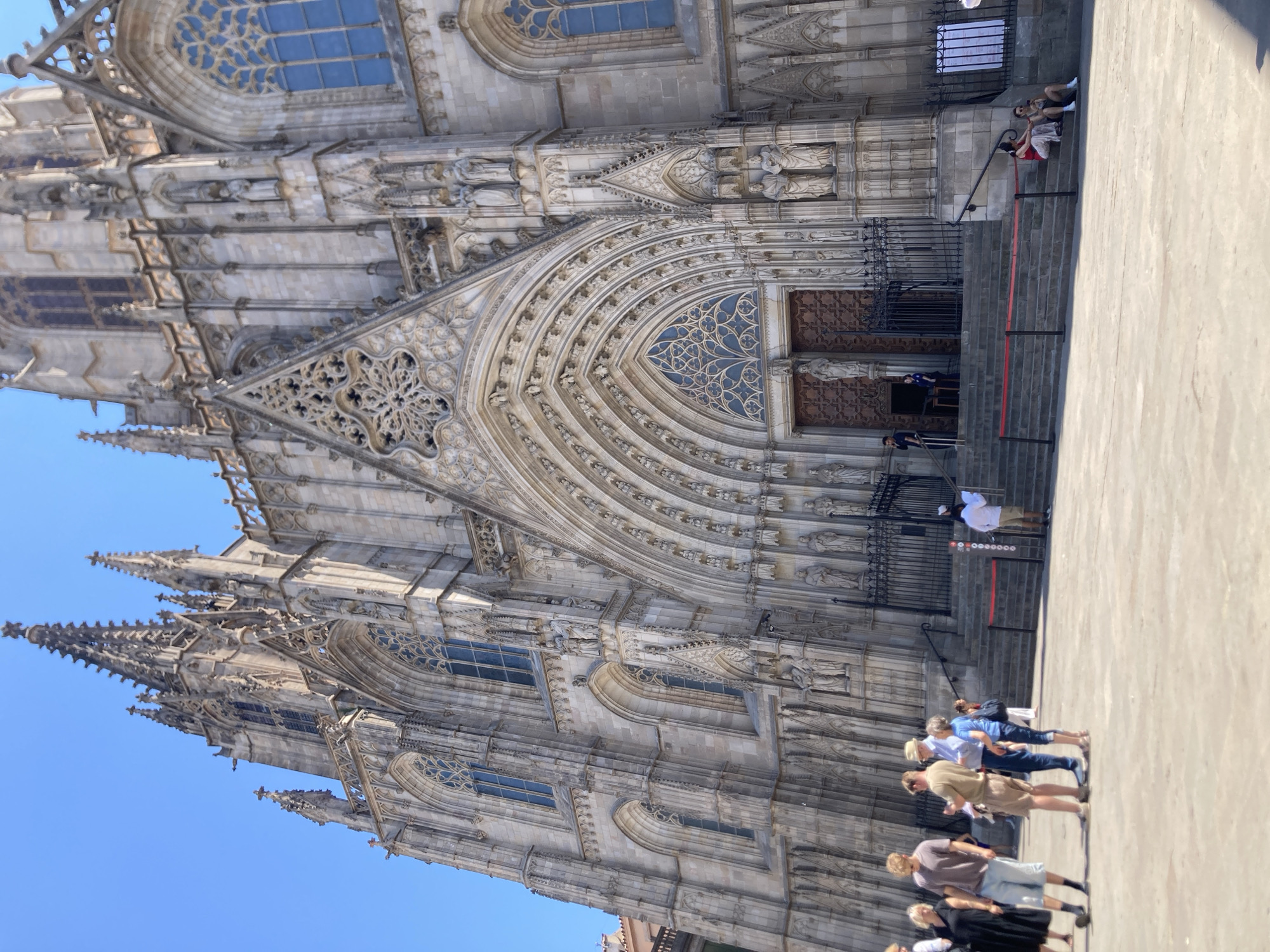 The image shows the exterior of the Barcelona Cathedral (Catedral de Barcelona) in Barcelona, Spain, on a sunny day. The cathedral is a Gothic Roman Catholic cathedral with intricate stone carvings, spires, and large arched entrances. A group of people are standing and sitting on the plaza in front of the cathedral. On the left, a group of four people are standing. One woman in a black dress and sandals is on the far left. Next to her, a man wearing a grey t-shirt, blue shorts, and black socks is facing to the right. To his right, a man in a light brown t-shirt and beige shorts is facing forward. Next to him, a man in a blue shirt, dark pants, and a straw hat is facing forward. Further to the right and further back, several other people are visible, some sitting on the steps of the cathedral and others standing near the entrance. The plaza is paved with light-colored stone. The sky is clear and blue.