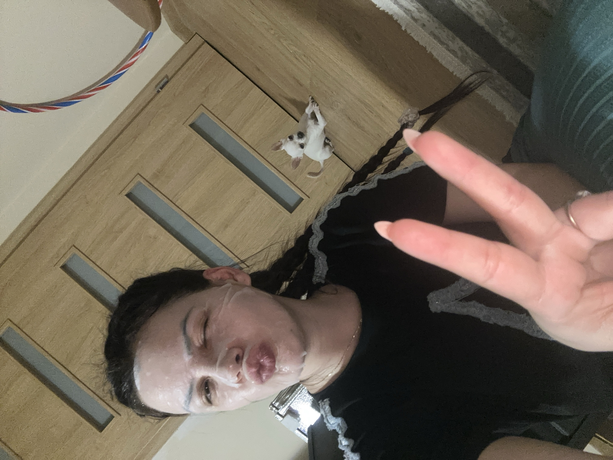 The image shows a woman with a facial sheet mask lying on her back indoors. She's wearing a black shirt and holding up a peace sign with her right hand. A small, white and black dog is lying on the wooden surface next to her. There is a hula hoop visible in the top left corner. The location is likely indoors, but there's no information in the image to specify the city of Chubra, Bulgaria.