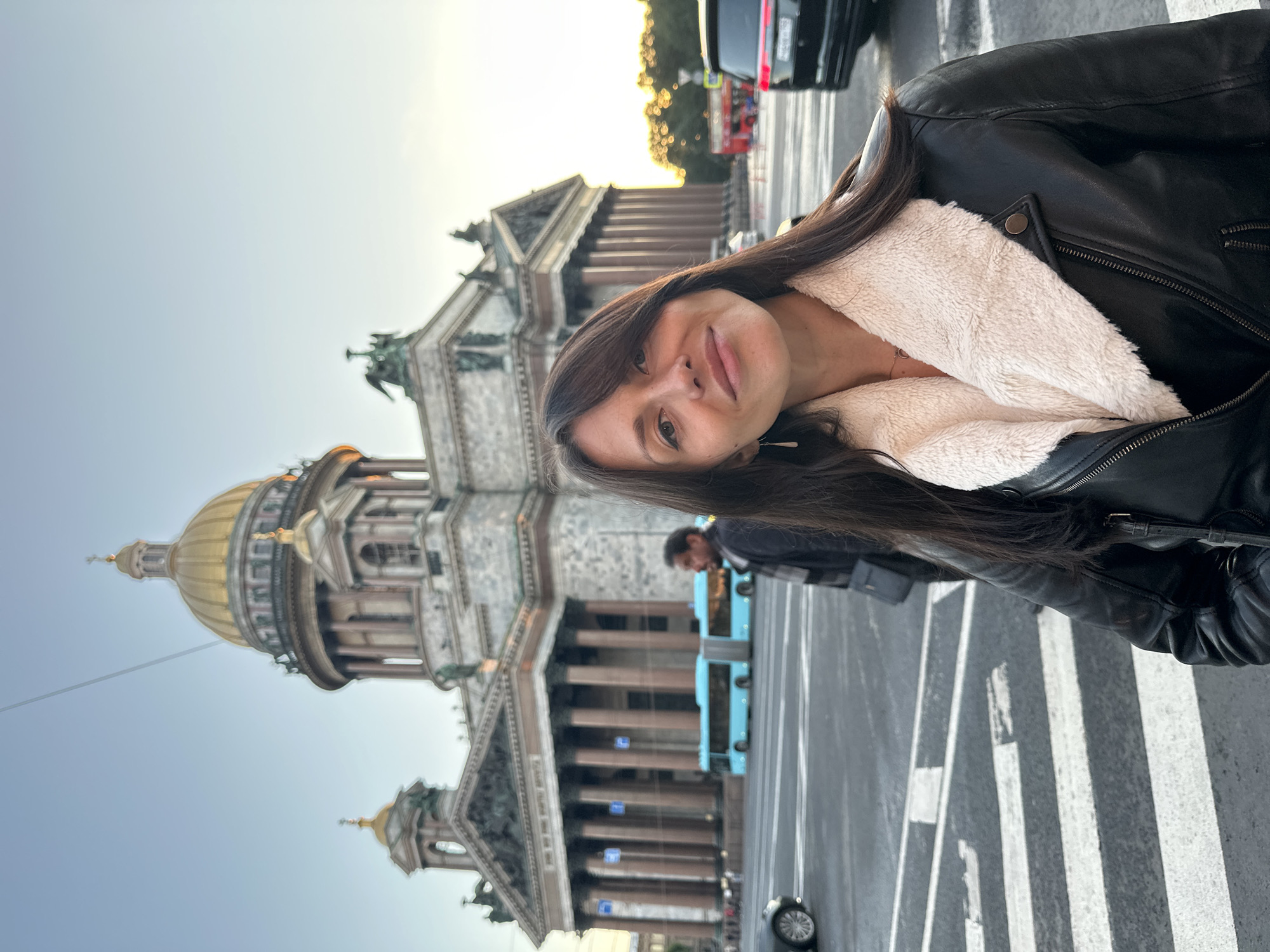 This is a medium shot portrait photograph taken outdoors in front of Saint Isaac's Cathedral in Saint Petersburg, Russia. A woman with long, dark hair is the primary subject, positioned in the foreground and slightly to the right of the frame. She is wearing a black leather jacket with a fluffy cream-colored collar and a dark top underneath. Her gaze is directed towards the camera with a gentle smile.
In the background, Saint Isaac's Cathedral stands prominently, characterized by its grand classical architecture, large columns, and a prominent golden dome. To the left of the cathedral, a bright blue articulated bus is visible on a street, indicating a public transportation system. A pedestrian crossing with white stripes is in the immediate foreground, suggesting the woman is standing on or near a street intersection. Other elements in the background include other vehicles, people, and trees, suggesting a busy urban environment. The lighting suggests it is either early morning or late afternoon, as the sky is a pale blue and the light has a warm quality.