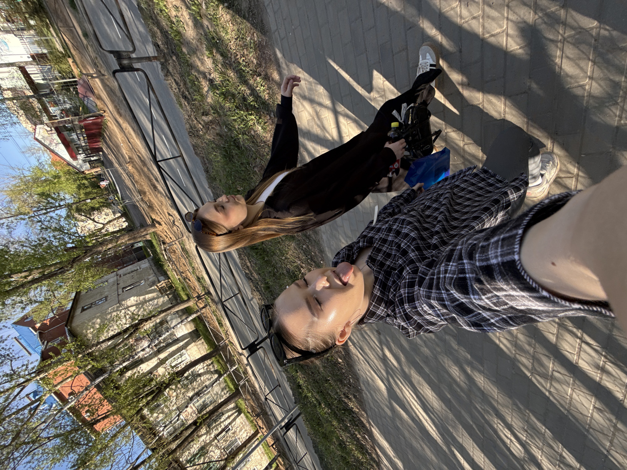 Two young women are standing on a paved walkway. The walkway runs adjacent to a road behind a metal fence. Buildings and trees are visible in the background. The sky is partially visible and is blue. The photograph appears to be a selfie, taken by the woman in the foreground wearing a black and white plaid shirt. She is sticking her tongue out. The other woman is standing behind her and is wearing a long black coat over a white top, and black pants. She has long blonde hair and sunglasses on her head. The location is Chaikovsky, Russia.