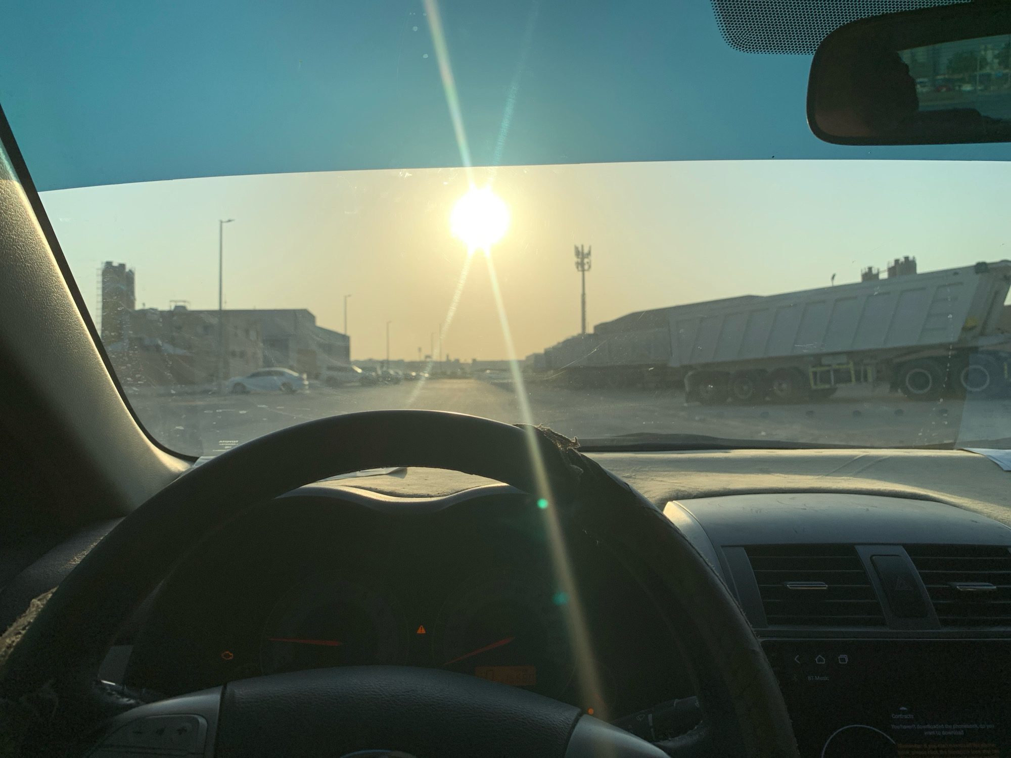 The image shows a view from inside a car in Musaffah, United Arab Emirates. The sun is low in the sky, casting a bright glare through the windshield. In the distance, there are buildings under construction, streetlights, and a large truck. The interior of the car is visible, including the steering wheel and dashboard. The overall impression is one of a developing urban or industrial area during sunset.