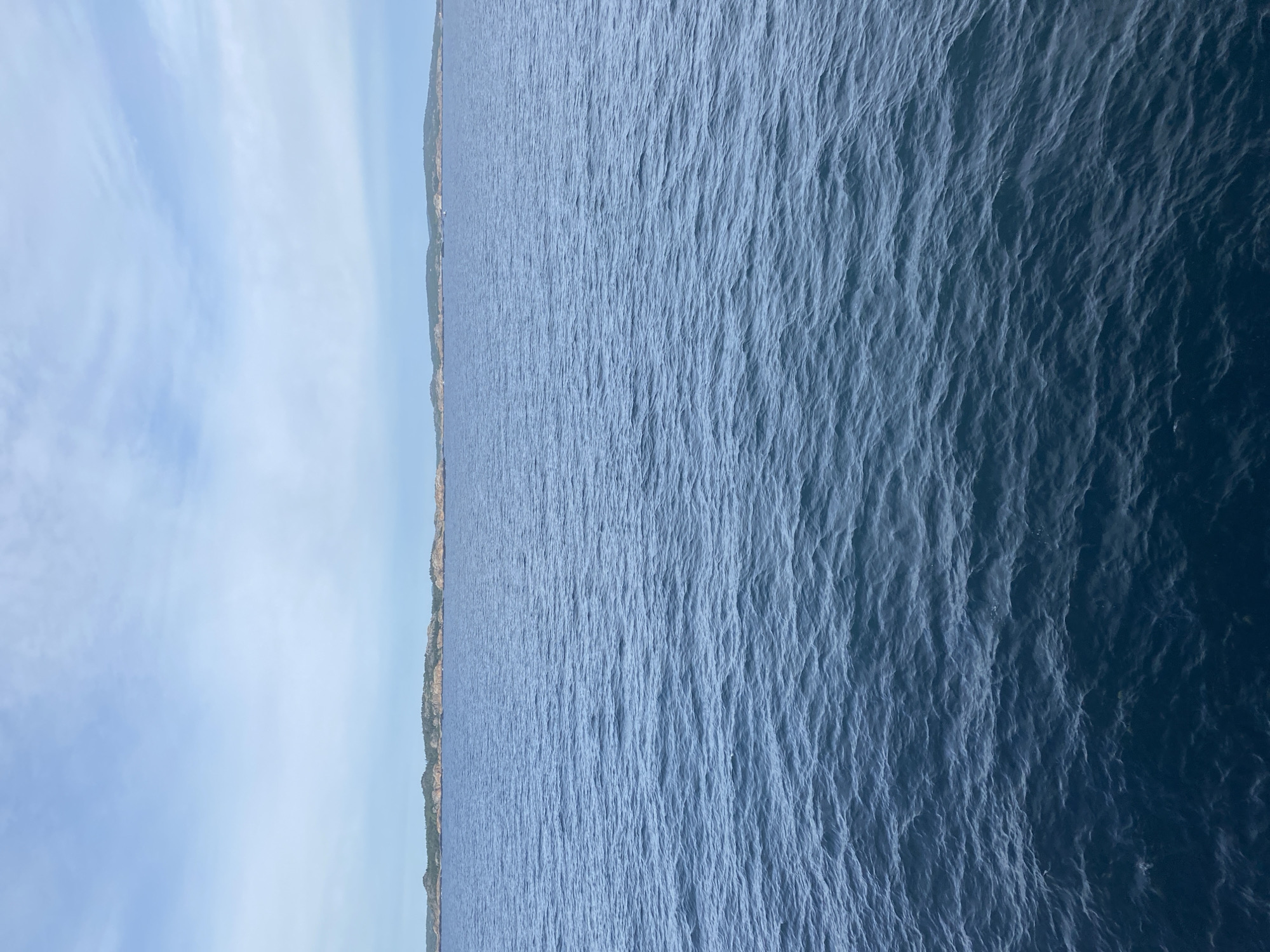 This is an aerial, landscape-oriented video clip shot at Stavern, Norway. The scene is dominated by the expansive blue sea, stretching across the majority of the frame. Gentle ripples and waves create a textured surface on the water, which appears to be a deep, rich blue. To the left of the frame, a sliver of coastline is visible, characterized by rugged terrain and green vegetation. Beyond the coastline, a clear blue sky occupies the upper portion of the image, with faint, wispy clouds scattered across it. The overall impression is one of a serene and vast natural landscape, likely captured on a clear day. The video is oriented vertically, suggesting it was filmed while looking down from a higher vantage point such as a drone or helicopter. There is no discernible activity or text within the frame.