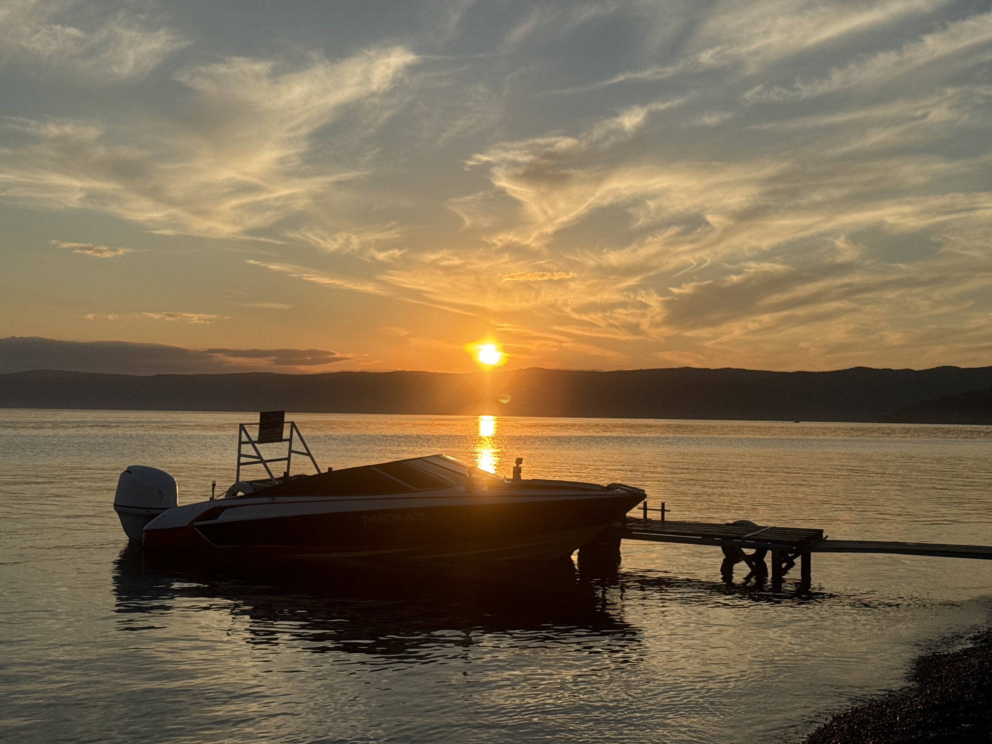 The image depicts a serene sunset scene on a body of water, likely Lake Baikal, near the village of Listvyanka, Russia. The sun, a bright orange orb, is setting on the horizon, casting a warm glow across the sky and water. Wispy clouds, tinged with yellow and orange, stretch across the sky. In the foreground, a speed boat is docked at a wooden pier. The boat is dark with a red stripe along its side, and an outboard motor is visible at the stern. The water is calm, reflecting the colors of the sunset. The shoreline in the distance is silhouetted against the sky, with mountains forming a dark band. The overall atmosphere is peaceful and tranquil, evoking a sense of natural beauty and quietude.