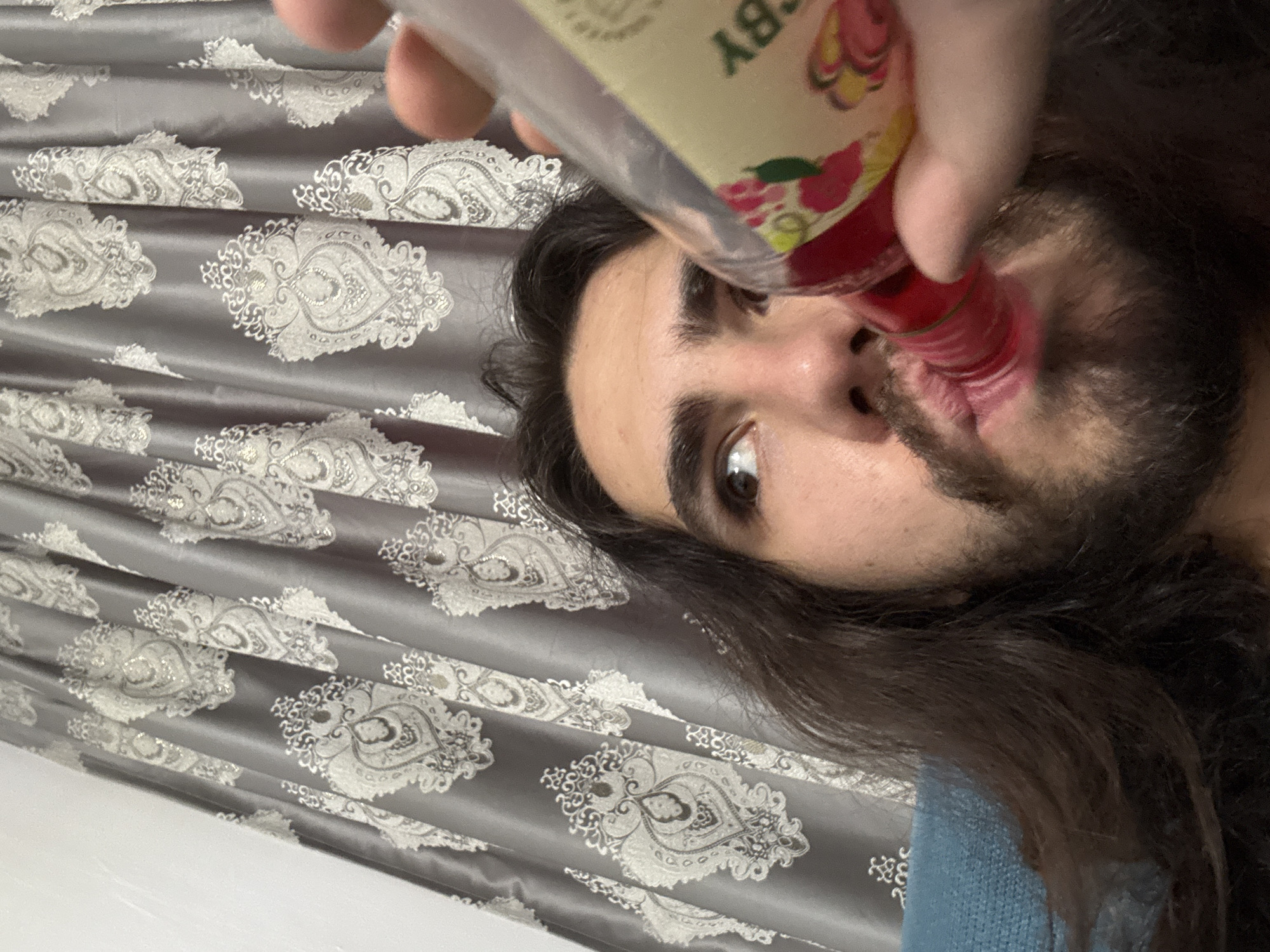 A close-up, eye-level, over-the-shoulder shot shows a young man with long, dark hair and a beard drinking from a bottle of red liquid. His head is tilted back, and he is looking to his right. The bottle is held in his right hand, with his thumb and forefinger visible. The background is a gray curtain with a white damask pattern. The scene appears to be indoors, possibly in a home.