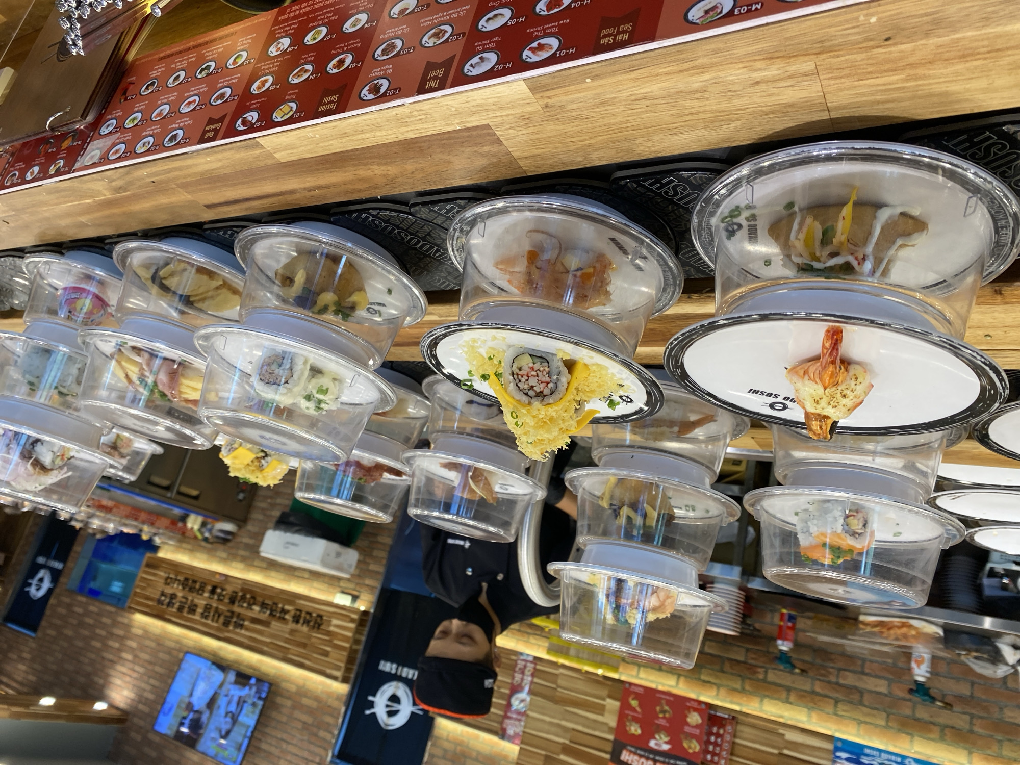 The image shows a conveyor belt sushi restaurant, likely located in Nha Trang Ward, Vietnam, given the Vietnamese text visible on the menu boards at the bottom. In the foreground, multiple plates of sushi are presented on clear plastic domes, stacked on a wooden counter. The sushi plates are circular, some with black rims. Several sushi rolls are visible, including a California roll with shredded yellow topping and a piece of shrimp. One plate features a large, cooked shrimp. Another plate holds a fried pastry-like item topped with white sauce and colorful julienned vegetables. In the middle ground, a person wearing a black chef's uniform, a black hat, and a black face mask is working behind the counter, arranging food items. Behind the chef, shelves hold more sushi dishes, and kitchen equipment such as torches are visible. The background reveals a brick wall with wooden accents and screens displaying images and text, including Vietnamese script. The overall environment suggests a busy, operational food establishment.