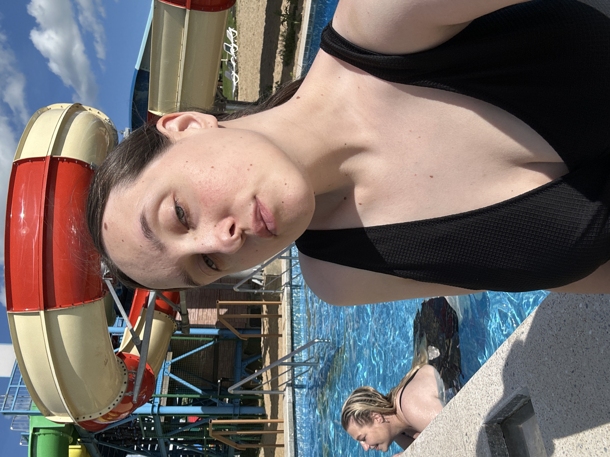 A woman is taking a selfie in a swimming pool at a water park. She is wearing a black swimsuit and has brown hair. In the background, there are water slides and lounge chairs. Another woman with blonde hair is in the pool. The sky is blue with some clouds. The location is the city of Yubileyny, Russia.