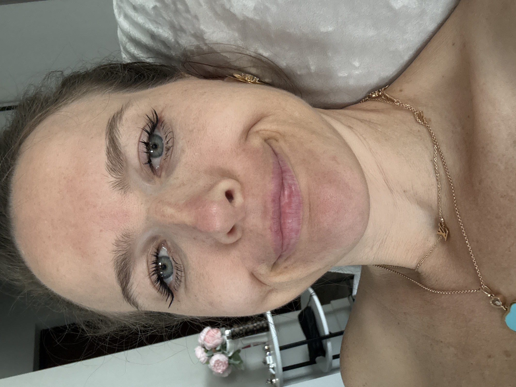 This image is a close-up, rotated portrait of a woman taken indoors. The woman's head is tilted to the right in the frame, and she is looking directly at the camera with a soft, pleasant smile. She has light skin, blue-grey eyes, and light brown hair, with visible mascara on her eyelashes.
She is adorned with gold-colored jewelry. A small earring is visible on her right ear, bearing the clear inscription "CRUZ". Around her neck, she wears a delicate gold-colored chain necklace. This necklace features a small, stylized initial charm (resembling an 'N' or 'M') near her collarbone, and a larger, light blue, possibly heart-shaped or cloud-shaped pendant lower on her chest.
The background behind her head and shoulders consists of soft, light-colored fabric, likely a pillow or bedding, suggesting a relaxed, personal setting such as a bedroom. In the lower left corner of the frame, a small decorative bouquet of light pink artificial flowers, possibly roses, is visible. To the lower right, there is a partial view of a metallic or white-framed object, possibly furniture.
The woman's expression conveys a calm and relaxed mood. The lighting appears to be natural, consistent with daytime. While the specific location cues are not evident within the image itself, the provided context for this media file is Dolgoprudny, Russia.