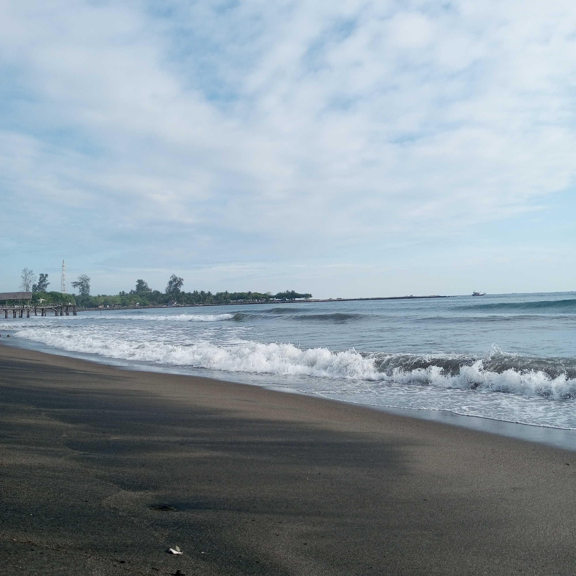 This media file captures a serene coastal landscape in Lhokseumawe, Indonesia. The foreground features a wide expanse of dark, fine-grained sand, characteristic of volcanic beaches, with small ripple patterns and a single white shell or piece of debris visible towards the bottom right. Gentle waves, topped with white foam, are seen breaking onto the shore and receding, creating a dynamic edge between the sand and the sea.
In the midground, the ocean extends outwards, displaying varying shades of blue and gray under a soft, diffused light. Further out, the water appears calmer, with subtle swells. On the far left, a wooden pier or jetty extends into the water, ending with a small covered structure. Behind the pier, a lush green coastline with various trees, including tall palms, is visible. A prominent, tall, lattice-like tower or antenna rises above the trees behind the pier. To the far right, a low-lying landmass or breakwater stretches across the horizon. A small boat or fishing vessel can be seen in the distance on the water, moving slowly near the horizon line towards the right.
The sky, occupying the upper half of the image, is a light, hazy blue with scattered, soft white clouds that obscure direct sunlight, suggesting an overcast or partly cloudy day. The overall lighting is even and soft, indicating it might be morning or late afternoon, rather than harsh midday. No discernible text is visible within the image. The scene conveys a sense of tranquility and natural beauty.