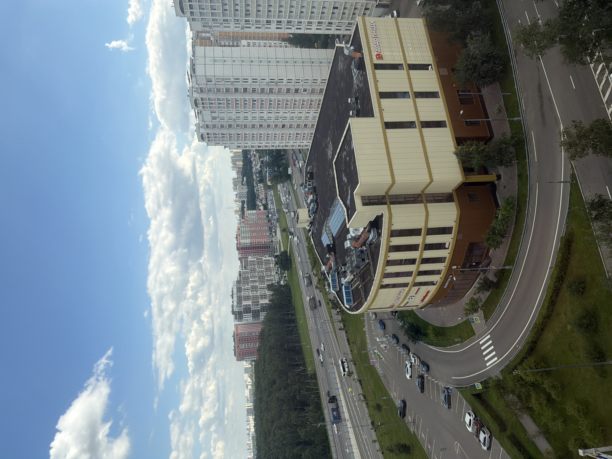 This image depicts a high-angle view of a multi-lane highway bordering a forest and a curved road leading to a light-colored, multi-story building with a brown base. The building has a prominent sign with Russian text and a circular logo. Several cars are parked in a lot adjacent to the curved road, and a few vehicles are visible on the highway, including trucks and buses. In the background, numerous tall, modern apartment buildings are scattered across the urban landscape, under a bright blue sky with scattered white clouds. The foreground includes a grassy area with trees and a lamppost.