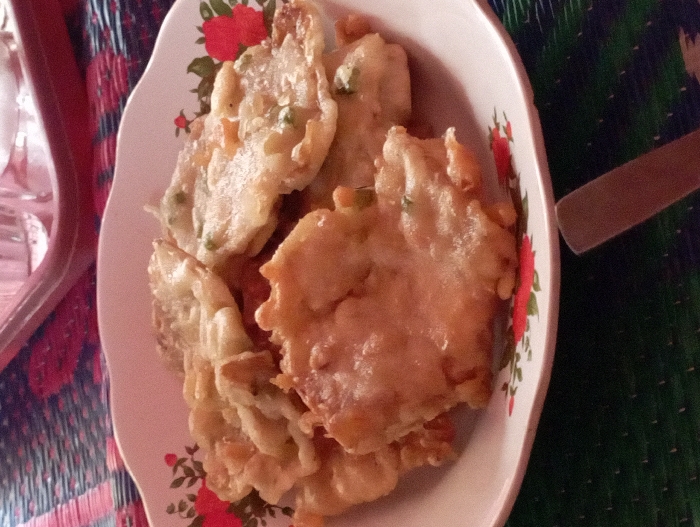 A close-up, overhead shot features a white plate filled with several pieces of golden-brown fried food, likely fritters, resting on a colorful woven mat. The plate has a distinctive red floral design bordering its rim. The fritters are irregularly shaped, with a crispy appearance, and some show visible specks of green ingredients, possibly chopped herbs or scallions, embedded within the batter.
The background consists of a woven mat, displaying patterns primarily in shades of green and red, suggesting a casual indoor setting, possibly a home. To the far left, a blurred portion of a clear glass or plastic container is partially visible. On the right side of the frame, the handle of a utensil, such as a spoon or knife, extends into view.
The scene depicts food prepared and ready for consumption. No people are visible, and there is no discernible text within the image. The setting and type of food are consistent with a meal or snack in Pubian, Indonesia.