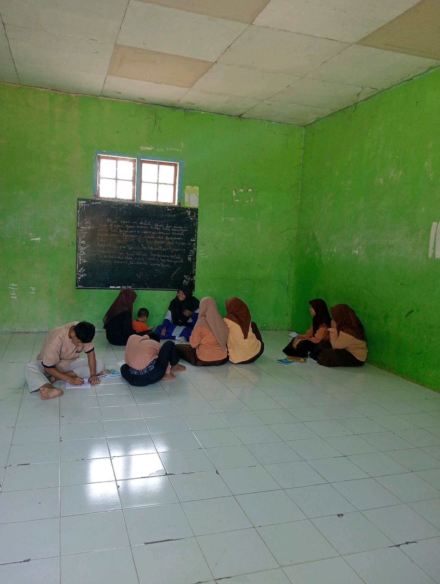 In a room with green walls and a tiled floor in Pagerungan Kecil, Indonesia, a group of people are sitting on the floor. A young man on the left is writing in a notebook. To his right, two women in hijabs are facing away from the camera, one with her back to the viewer and the other facing right. Further right, a woman in a dark hijab is sitting, and a young child in an orange shirt is beside her. Another woman in a tan hijab is next to them. On the far right, two women are seated, both wearing hijabs and tan uniforms. A blackboard with writing is mounted on the green wall behind them, and a window with four panes is above it. The room appears to be a classroom or community space.