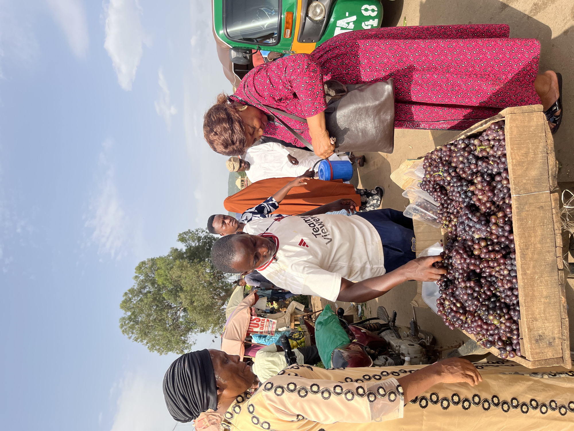 The image depicts a busy outdoor market scene in Rafin Par, Nigeria. The primary subjects are several people, seemingly engaged in commerce. In the foreground, a woman wearing a light-colored, ornately decorated dress and a black headscarf is looking upwards, her mouth slightly open as if speaking. To her right, a man in a white t-shirt with red lettering that reads "Team Viewer" is reaching towards a large wooden crate overflowing with dark purple grapes. Another person, partially visible behind him, wears a white robe and a cap. Further back, a woman in a vibrant pink dress with a floral pattern is bent over, looking towards the ground, with a brown leather bag slung over her shoulder. Behind her, a green and yellow three-wheeled auto-rickshaw is parked, suggesting transportation is readily available. The background shows a clear blue sky with scattered white clouds, indicating a sunny day. The ground appears to be dirt or unpaved road, typical of many Nigerian markets. The overall atmosphere suggests an active marketplace with transactions taking place. The lighting and clear sky suggest it is daytime, likely late morning or early afternoon.