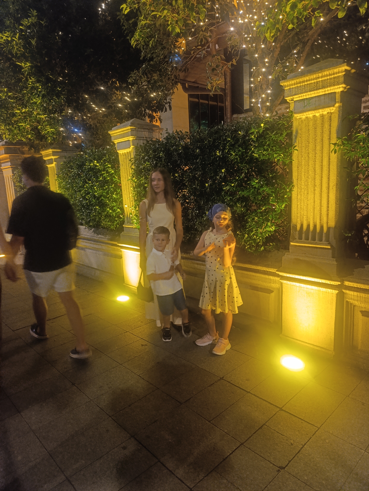 The image depicts a nighttime scene on a paved walkway, likely in Batumi, Georgia, illuminated by warm yellow spotlights and decorative string lights in the trees. In the foreground, a person is walking away from the camera, wearing a black t-shirt and white shorts. To their right, a woman in a light-colored dress stands with a young boy in a white polo shirt and blue shorts. Beside them, a young girl in a yellow polka-dot dress and a blue headscarf is posing. Behind the group is a dense green hedge, and ornate yellow pillars with fluted details are visible, suggesting a public space or promenade. The atmosphere is festive and suggests an outdoor gathering or stroll.