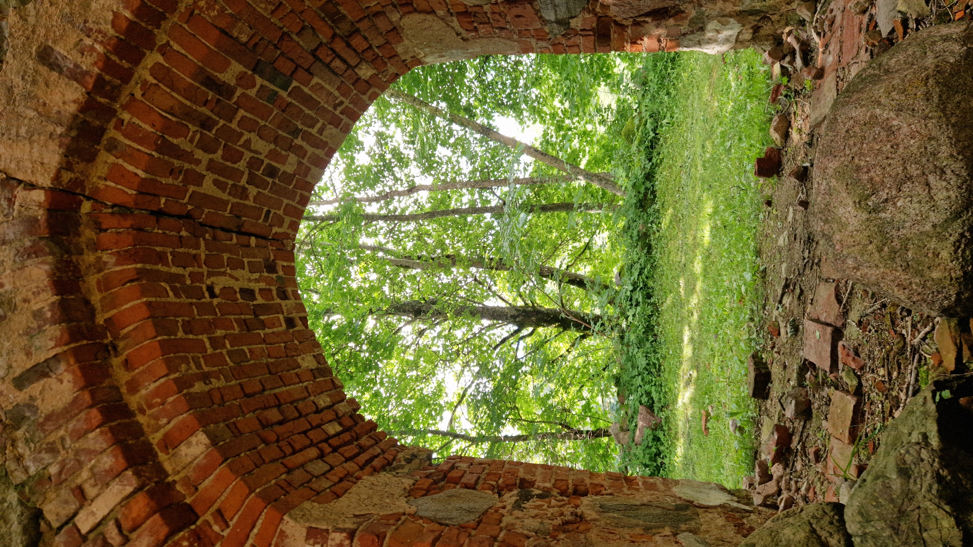 The image depicts an arched opening in a ruined brick structure, looking out onto a lush, green outdoor scene. The foreground is dominated by weathered red bricks, forming the archway, with some rough stones and rubble visible along the edges of the opening. The bricks show signs of age and decay, with visible mortar and some cracks.
Through the arch, a bright, sunlit area of green grass and dense foliage is visible. Several trees with full, green canopies are prominent, their branches reaching upwards and outwards. The sunlight filters through the leaves, creating dappled patterns on the ground. The scene suggests a warm, possibly summer day. The overall impression is one of nature reclaiming an old, man-made structure. There are no people or discernible text visible in the image. The location is contextually identified as Embūte, Latvia.