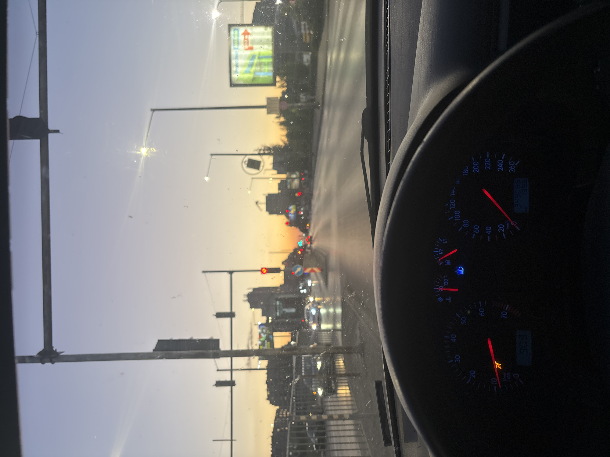 The image shows a view from inside a car, likely the driver's seat, while on a road in Sofia, Bulgaria. The dashboard is visible, displaying a speedometer reading around 20 km/h and an RPM gauge indicating approximately 909 RPM. Outside the windshield, the scene is dominated by an urban landscape during what appears to be either sunset or sunrise, given the warm color gradient in the sky. Streetlights and a large billboard are present, along with typical city buildings and traffic lights, some of which are red. The road seems busy with several cars.