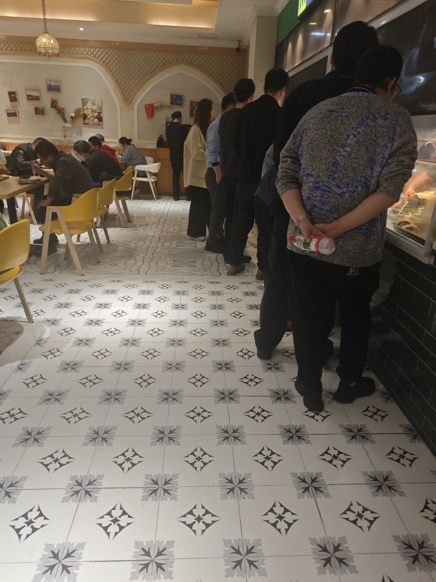 The image shows a line of people waiting at a food counter in a restaurant or cafeteria, likely in the city of Chongqing High-tech Zone, China. The customers are primarily facing away from the camera, focusing on the food display. They are dressed in casual attire. One person in the foreground is holding a water bottle behind their back. The restaurant features patterned tile flooring, yellow chairs, and decorative archways. Several people are seated at tables in the background, eating. There is a light fixture hanging from the ceiling.