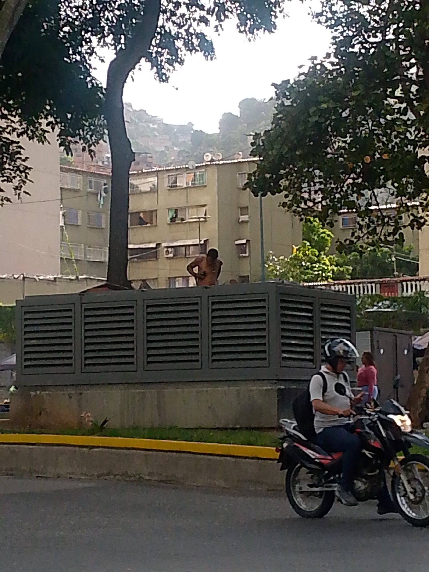 The image depicts an urban scene in Caracas, Venezuela. A shirtless man stands atop a large, dark gray, louvered structure next to a tree on a median strip. Behind the structure are residential buildings on a hillside. A person wearing a helmet and backpack is riding a motorcycle on the street in the foreground. A young girl is walking nearby on the sidewalk with another person, near what appears to be makeshift market tents with merchandise. The sky is overcast.