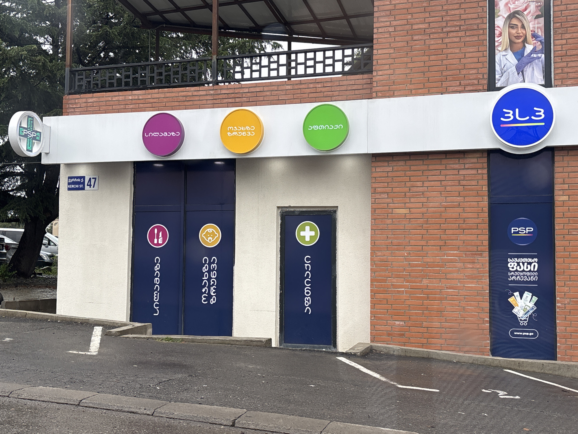 In Tbilisi, Georgia, on Kerchi Street, a white building with a brick facade features a pharmacy storefront. The facade has several circular signs with Georgian text and symbols. The left side of the building has a white PSP pharmacy logo. To the right are three circular signs in purple, yellow, and green, each with Georgian text. Below these signs are three dark blue doors. The left door has a symbol of a toothbrush and toothpaste. The middle door has a baby's face. The right door has a green cross. Further to the right, a large vertical blue sign with the PSP logo is mounted on the brick wall, and below it, more Georgian text and an illustration of money in a shopping cart. An advertisement for a woman is displayed in a frame above the blue PSP sign. The scene is viewed from a slightly elevated asphalted area that appears to be a parking lot, with white lines marking parking spaces. A tree and partial view of a car are visible to the left of the building. The sky is overcast.