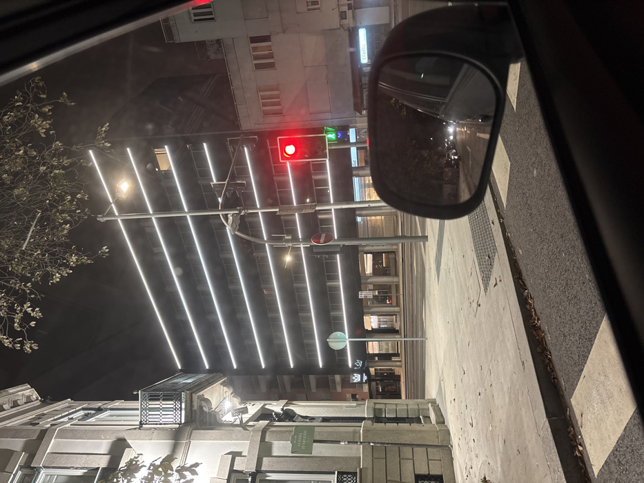 The image depicts a nighttime street scene from inside a stationary vehicle at the intersection of Rua Alegria Junqueiro and Rua de Dom Pedro V in Lordelo do Ouro e Massarelos, Porto, Portugal.
To the left, a multi-story building with a light-colored stone facade and an ornate black wrought-iron balcony is visible. A white street sign on its corner clearly reads "RUA ALEGRIA JUNQUEIRO." Directly ahead, a modern, dark-colored multi-story building dominates the background, characterized by multiple parallel horizontal white LED light strips illuminating its facade.
A tall street light pole stands centrally, supporting a vehicle traffic light displaying a solid red signal and a pedestrian traffic light displaying a green walking figure. Below these, a circular red 'no entry' sign is visible. The asphalt road features white pedestrian crossing stripes. The concrete sidewalk to the left is partially covered with scattered brown leaves.
The foreground includes the interior bottom frame of a vehicle window and the passenger-side exterior mirror. The mirror's reflection shows a dark street behind the camera's position, with the headlights of several distant vehicles visible. The overall scene is devoid of people.