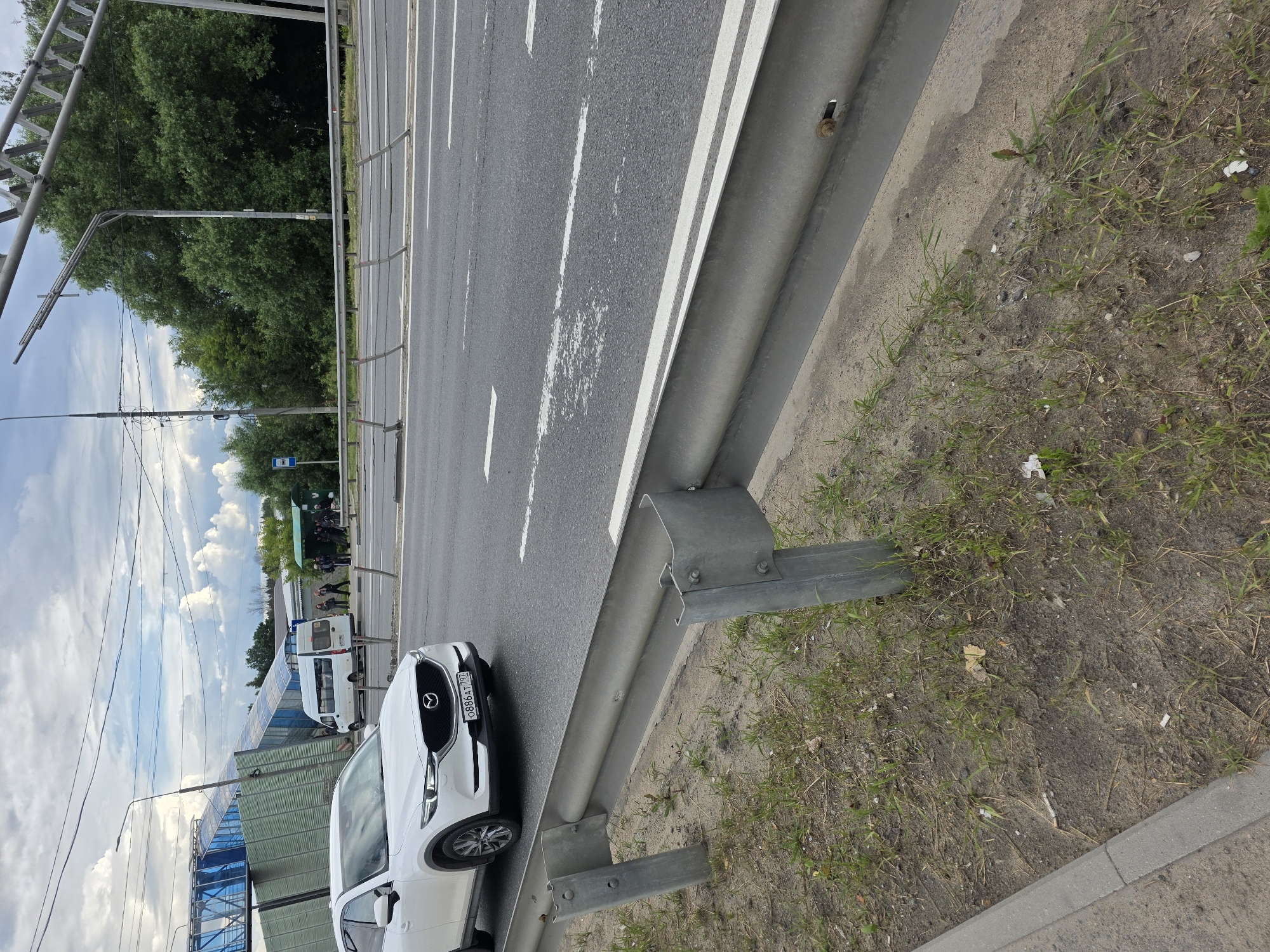 A white Mazda CX-5 is traveling on a multi-lane road. The car has a license plate that reads "0886AT 797". In the background, a white passenger van is parked near a bus stop. Several people are standing at the bus stop, some of whom appear to be waiting for transportation. A blue and green pedestrian overpass is visible in the distance, crossing over the road. The environment appears to be a suburban or semi-urban area with trees lining the side of the road. The sky is cloudy.