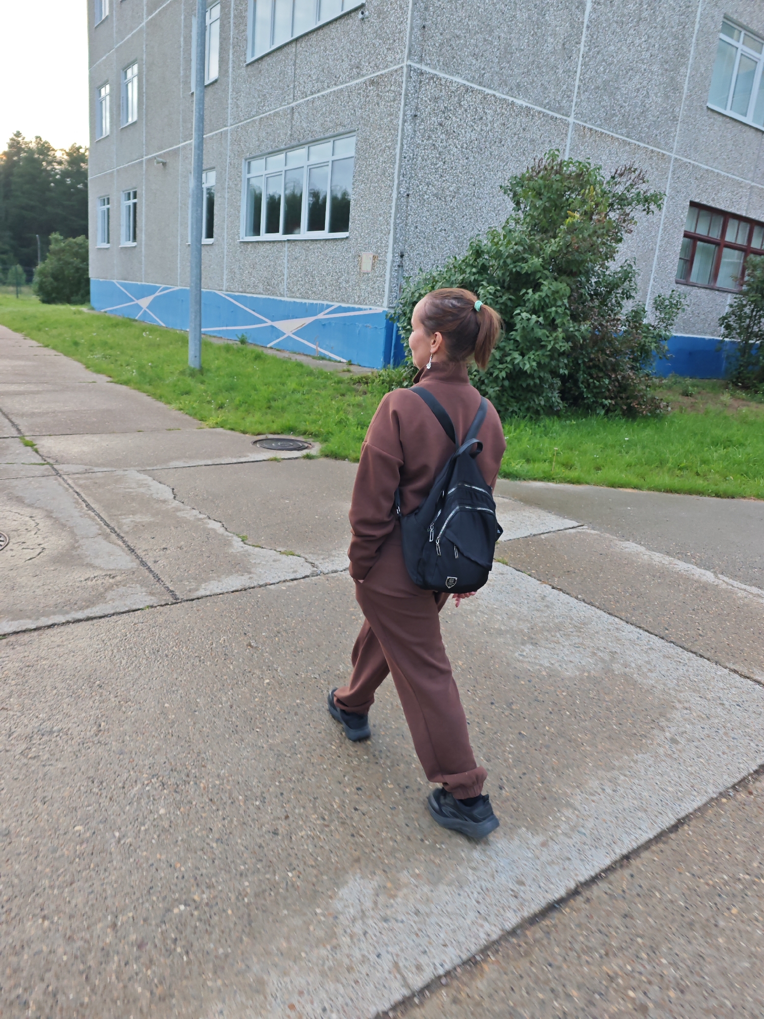 A person walks away from the camera on a concrete sidewalk next to a multi-story building. The person is wearing a brown tracksuit and a black backpack. The building has a grey, textured facade with blue and white decorative trim along the bottom. There is green grass and trees on the left side of the image and in front of the building. The scene is likely outdoors in a residential area.