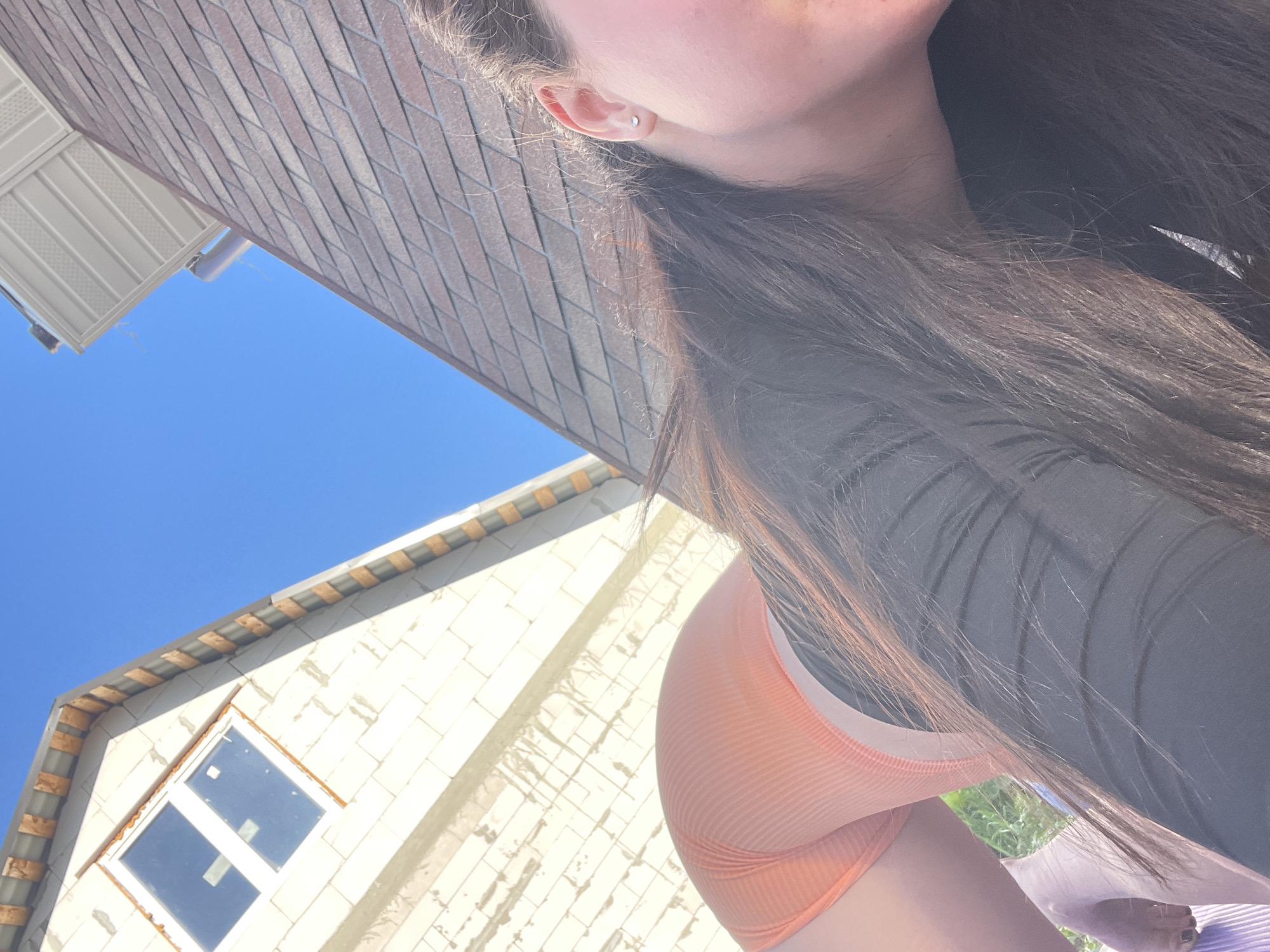 The image shows a woman from a low angle, with a building under construction in the background. The building is made of white blocks with a window. The sky is visible above the building. In the foreground, the woman is wearing a dark top and an orange ribbed garment. The location is likely in Мосметаллоконструкция Онт, Russia.
