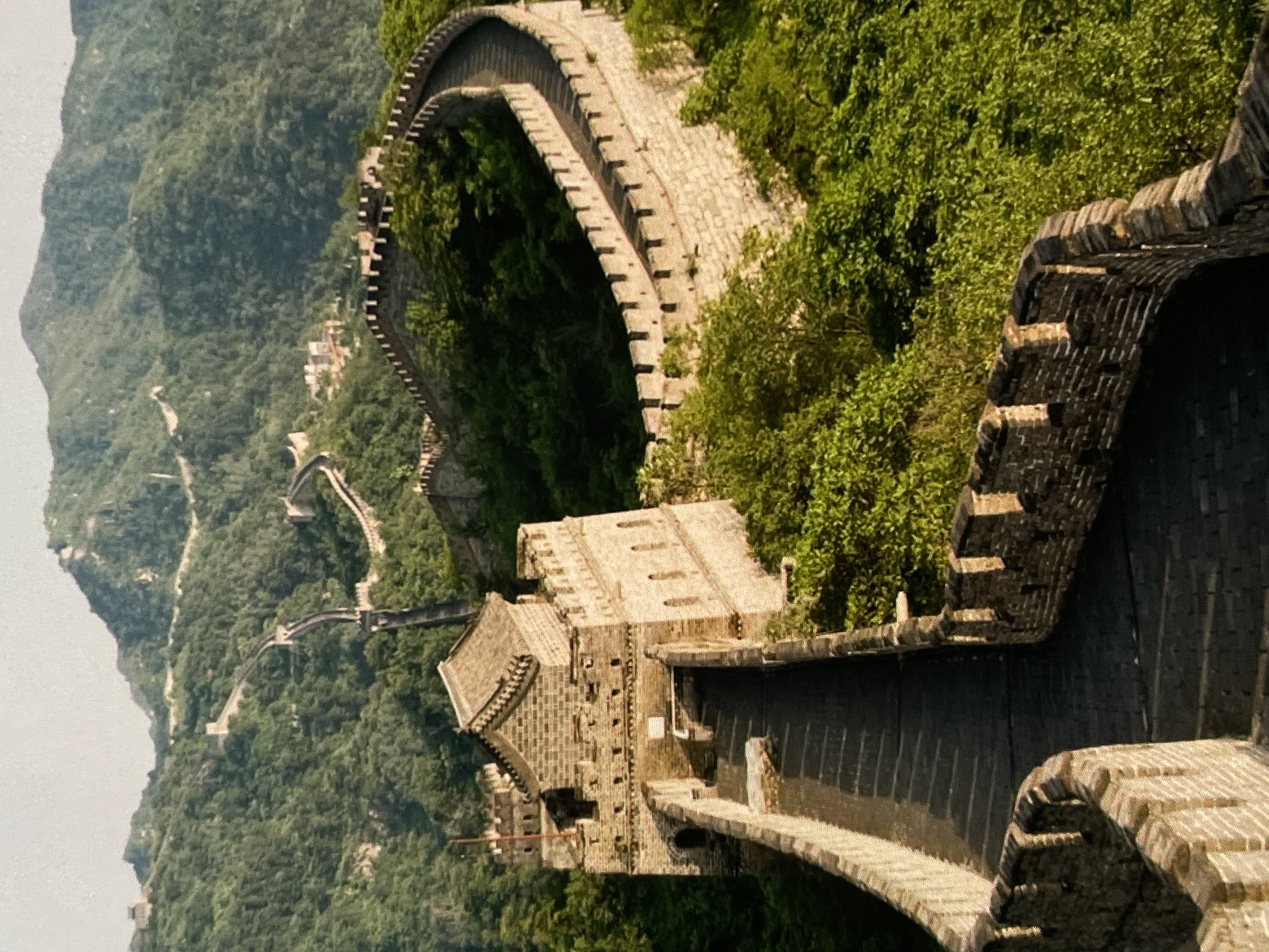 This is an aerial shot of a section of the Great Wall of China, winding through a lush, forested mountain landscape. The wall is constructed of stone and brick, with crenellated battlements visible along its length. Several watchtowers are integrated into the wall, one of which is prominent in the foreground, featuring a multi-tiered, tiled roof. The path of the wall snakes up and down the steep, green hills, disappearing into the distance where more sections are visible. The sun casts shadows on the wall and the surrounding trees, indicating it is likely morning or late afternoon. The environment is natural and undeveloped, with dense vegetation covering the mountains. There are no people or animals visible in the image. The image does not depict Harmanli, Bulgaria.