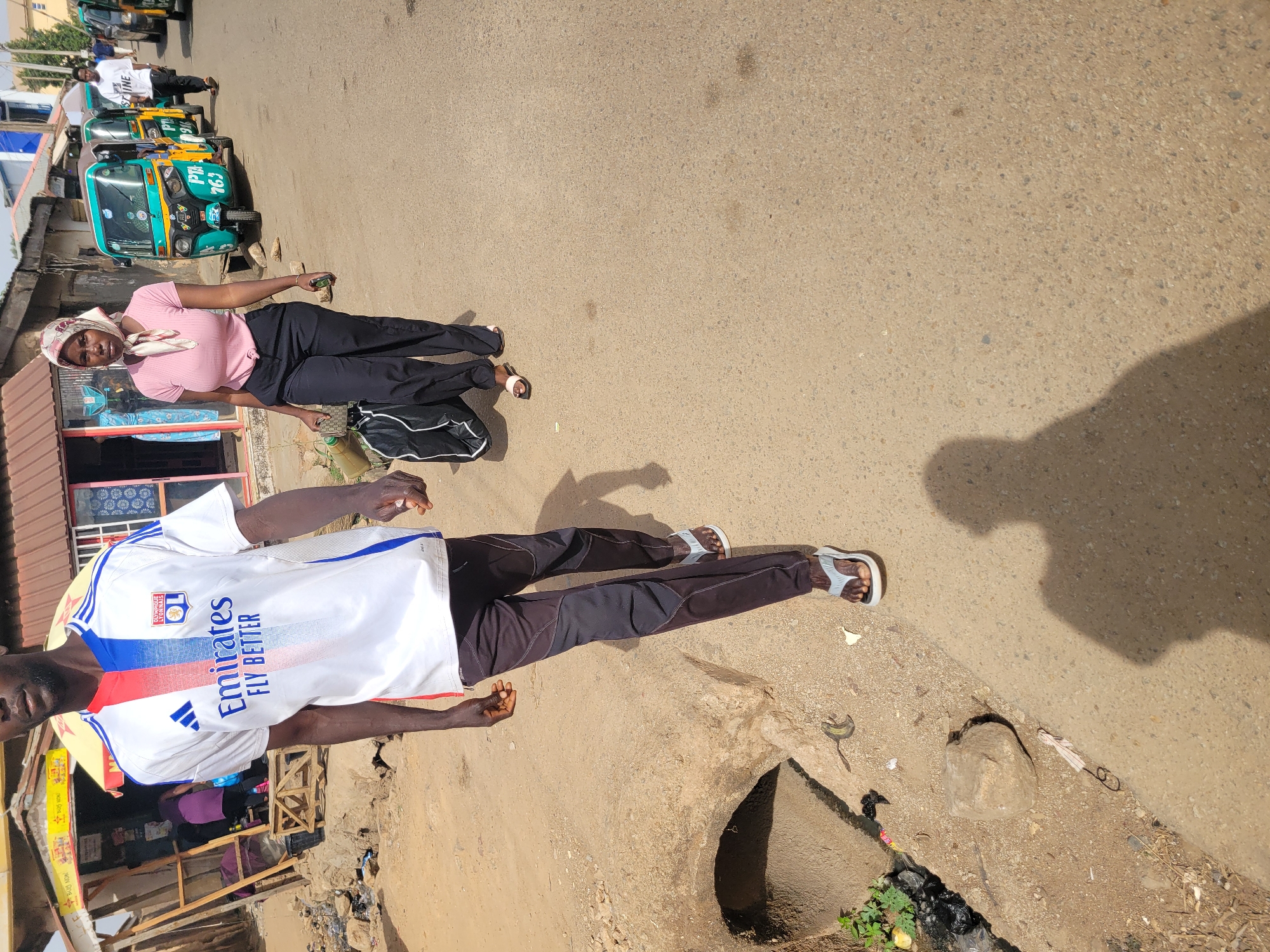 The image depicts a vibrant street scene in Jos, Nigeria, under bright daylight, evidenced by distinct shadows cast across the paved road. The time of day appears to be mid-morning or afternoon.
In the foreground, two individuals are prominent. On the left, a woman with dark skin wears a light pink t-shirt tied at the waist, black trousers, and light-colored sandals. A patterned headscarf covers her head. She holds a small, light green jerrycan or bottle in her right hand and has a black and white duffel bag resting at her feet. She is looking towards the camera.
To the right, a man with dark skin stands wearing a white short-sleeved football jersey with red and blue stripes and black athletic trousers paired with grey sandals. His jersey clearly displays the "OL" logo (Olympique Lyonnais) and the sponsor text "Emirates FLY BETTER". He is facing away from the camera's direct line of sight.
The street itself is made of tarmac or coarse asphalt, with some patches of dirt and litter visible, including an open drainage ditch or culvert in the lower right foreground. In the background, a row of green and yellow auto-rickshaws, locally known as Keke Napep, are parked. One tricycle prominently displays "PTB 763" on its front. To the far left, there are glimpses of small shops or market stalls with simple structures and various items on display, consistent with a local commercial area. Other people are visible further down the street among the tricycles. The scene conveys the bustling atmosphere of a Nigerian city street.