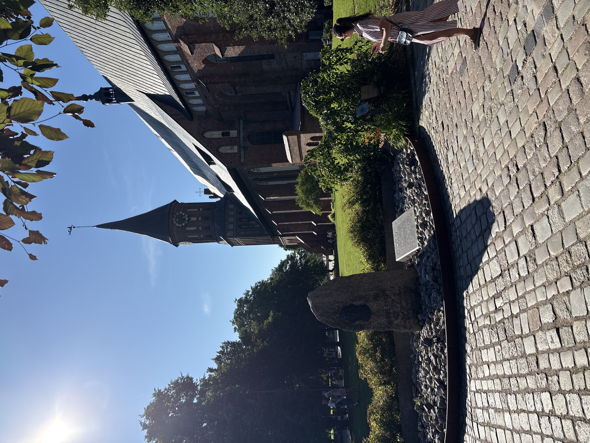 This image depicts a scene in front of the Kaliningrad Cathedral, located on Kant Island in Kaliningrad, Russia. The cathedral, a large brick Gothic structure with a prominent clock tower, dominates the background. In the foreground, a cobblestone pathway leads towards the cathedral. A large stone monument with a portrait and inscription is situated on the left, surrounded by gravel. A woman, wearing a striped dress and carrying a phone and a small bag, stands on the right side of the pathway, facing away from the camera. She appears to be looking at her phone or at the monument. Several trees and a lawn are visible behind the monument and to the left of the cathedral. The sky is clear and blue, with the sun casting shadows across the scene.