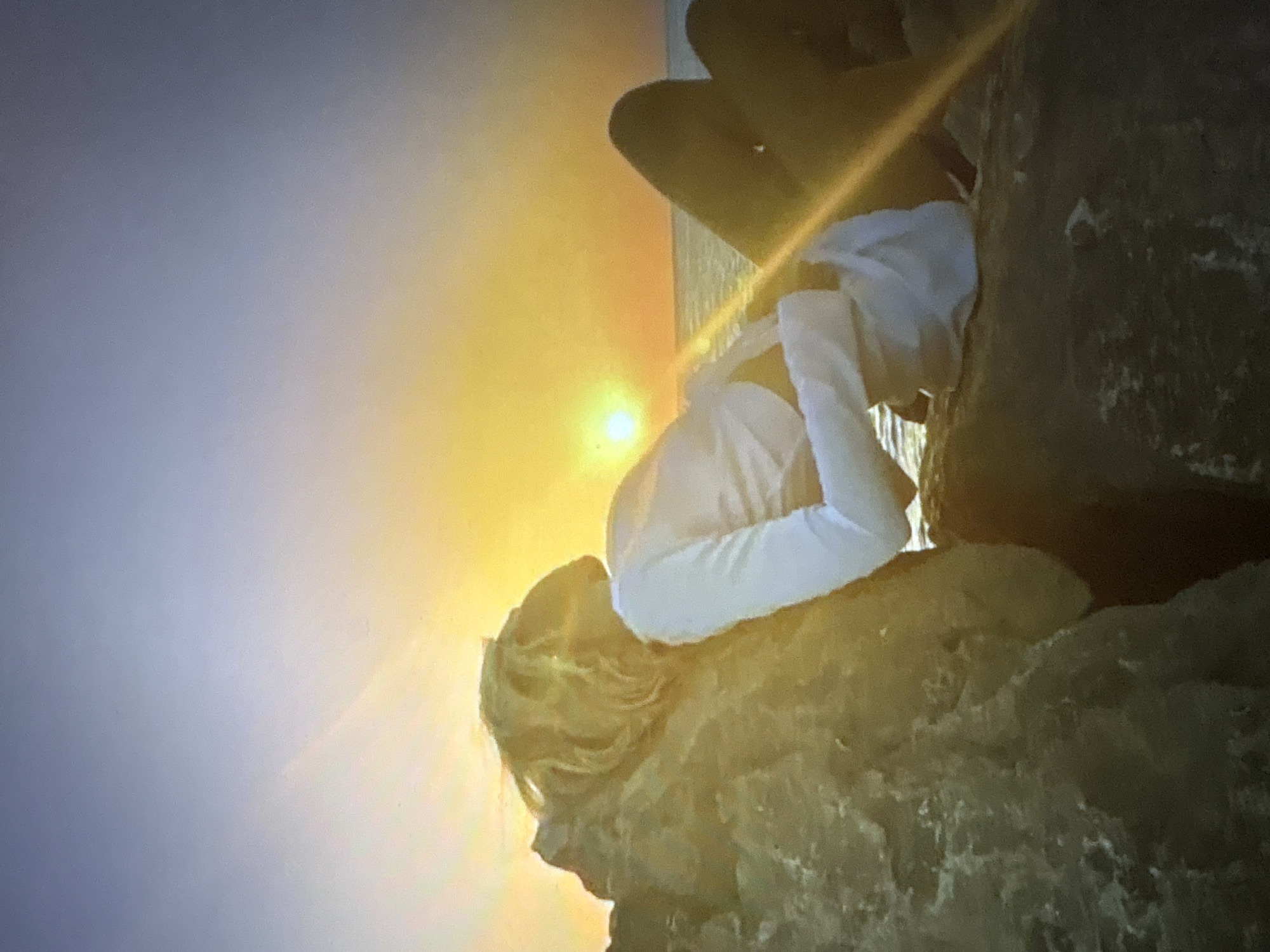 An outdoor, full-body shot captures a blonde person in a white long-sleeved shirt and dark bottoms reclined on rocks near the ocean. The person is positioned to the left of the frame, with their back to the camera, looking out at the sunset. The sun is low on the horizon, casting a warm orange and yellow glow across the sky and water. The ocean is visible in the background, with a faint horizon line. The rocks are rough and textured, providing a natural seating area. The scene is bathed in the soft light of dusk.