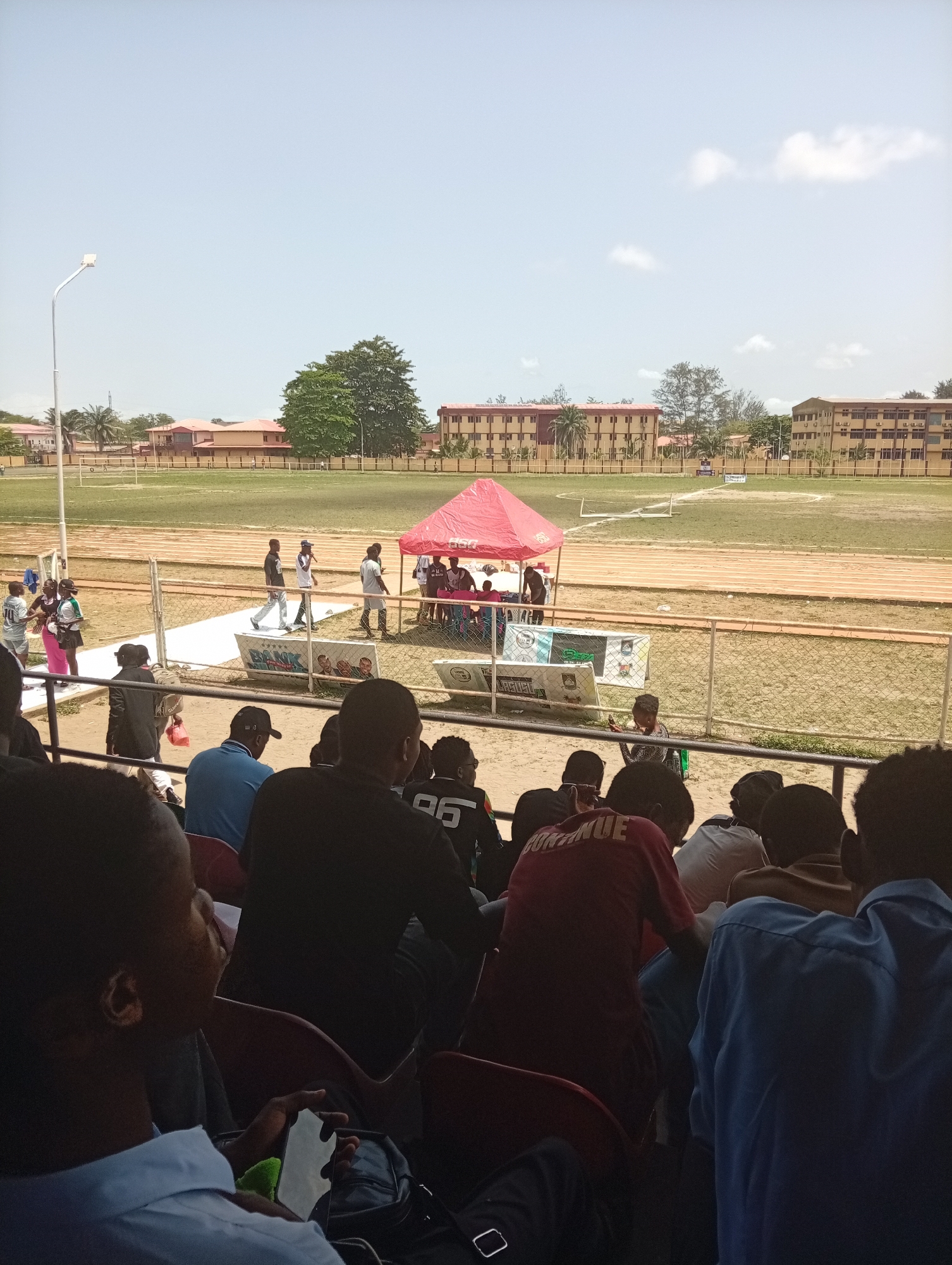 The image depicts a scene at what appears to be a sports stadium in Isasi, Nigeria. The foreground shows a section of seated spectators, most of whom are facing away from the camera. The middle ground features a running track and a grass field. A red tent is set up on the track, with people standing around it. A white banner or platform is placed in front of the tent, with logos and lettering visible. A wire fence separates the spectators from the track area. In the background, there are several multi-story buildings and trees. The sky is light blue with a few scattered clouds. A streetlight is visible on the left side of the field. People are walking on the track near the tent.