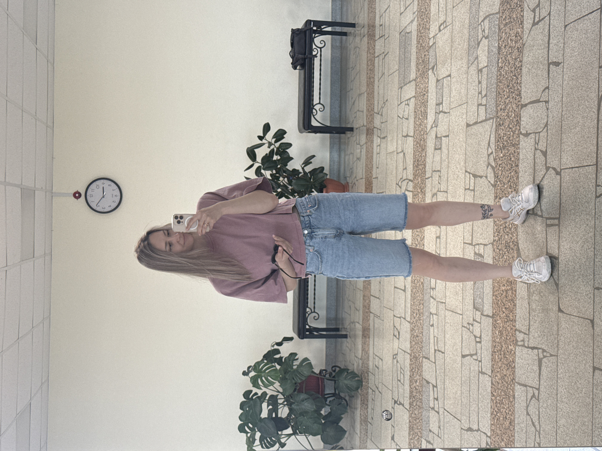 A full-length photograph depicts a woman indoors. She is fair-skinned, with long blonde hair, and is wearing a light purple t-shirt and blue denim shorts. She holds a smartphone in her right hand, presumably taking the picture.
The woman is positioned in what appears to be a public space, likely a lobby or waiting area, evidenced by the tiled flooring, neutral-colored walls, and the presence of a clock, potted plants, and metal benches.
The clock on the wall displays the time as approximately 2:50 PM.
The tiled floor features a pattern of grey and beige rectangular tiles.
Two potted Monstera plants are located near the lower portion of the frame. Behind the woman, two decorative metal benches.
The scene is generally well-lit, although shadows suggest the presence of direct light.
The presence of the Monstera deliciosa plants indicates a possible correlation with the city of Noviy Urengoy, Russia.