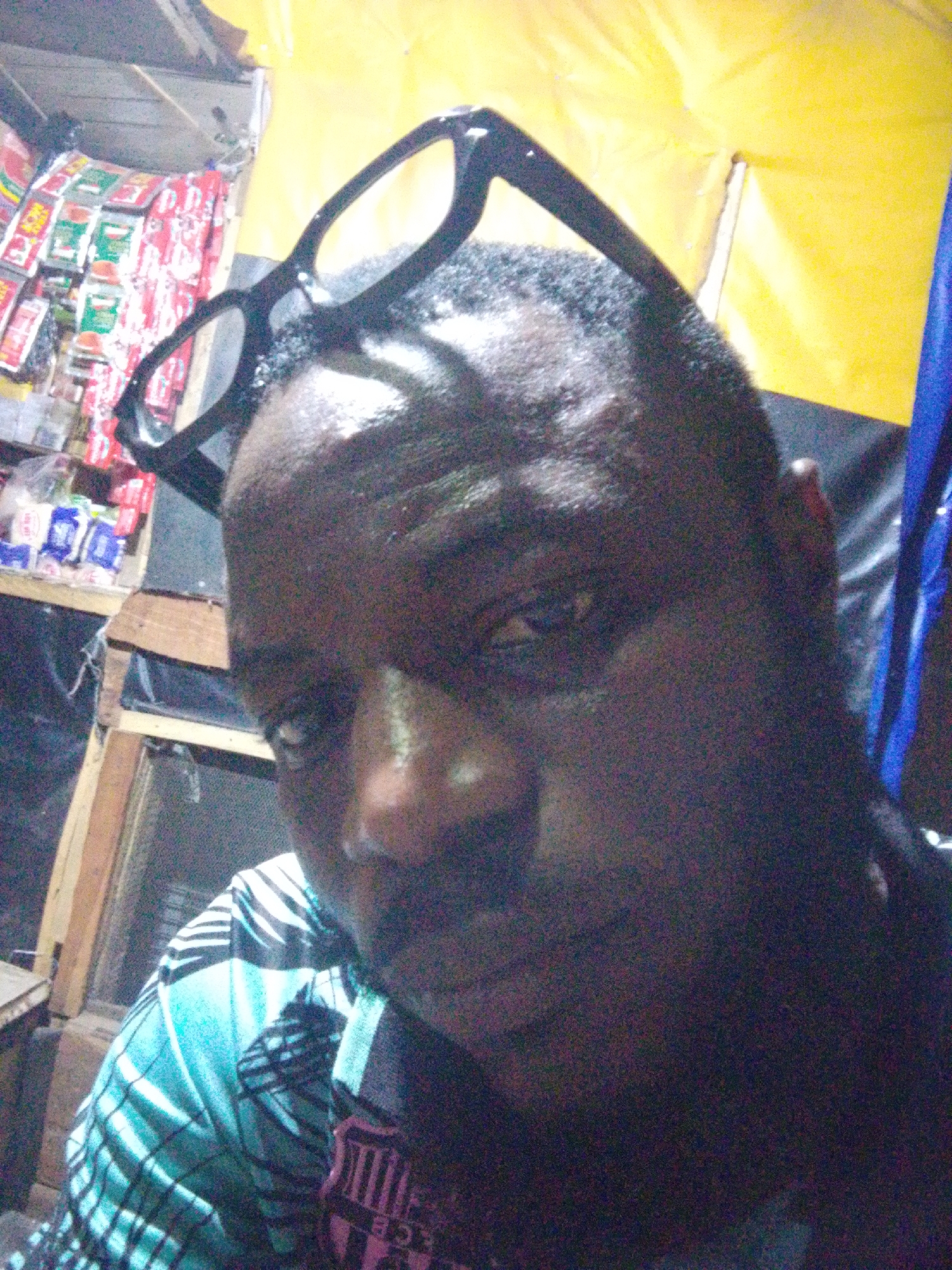 A close-up photograph shows a man of African descent wearing black-rimmed glasses perched on his forehead. The man has a short haircut and a beard. He is wearing a patterned shirt with light blue, white, and black stripes. In the background, shelves display packaged goods, indicating a store setting. The area behind the man appears to be draped with a yellow tarp. Based on the prompt's context, this location may be within the city of Ajaokuta, Nigeria. The photo does not show interactions or actions.